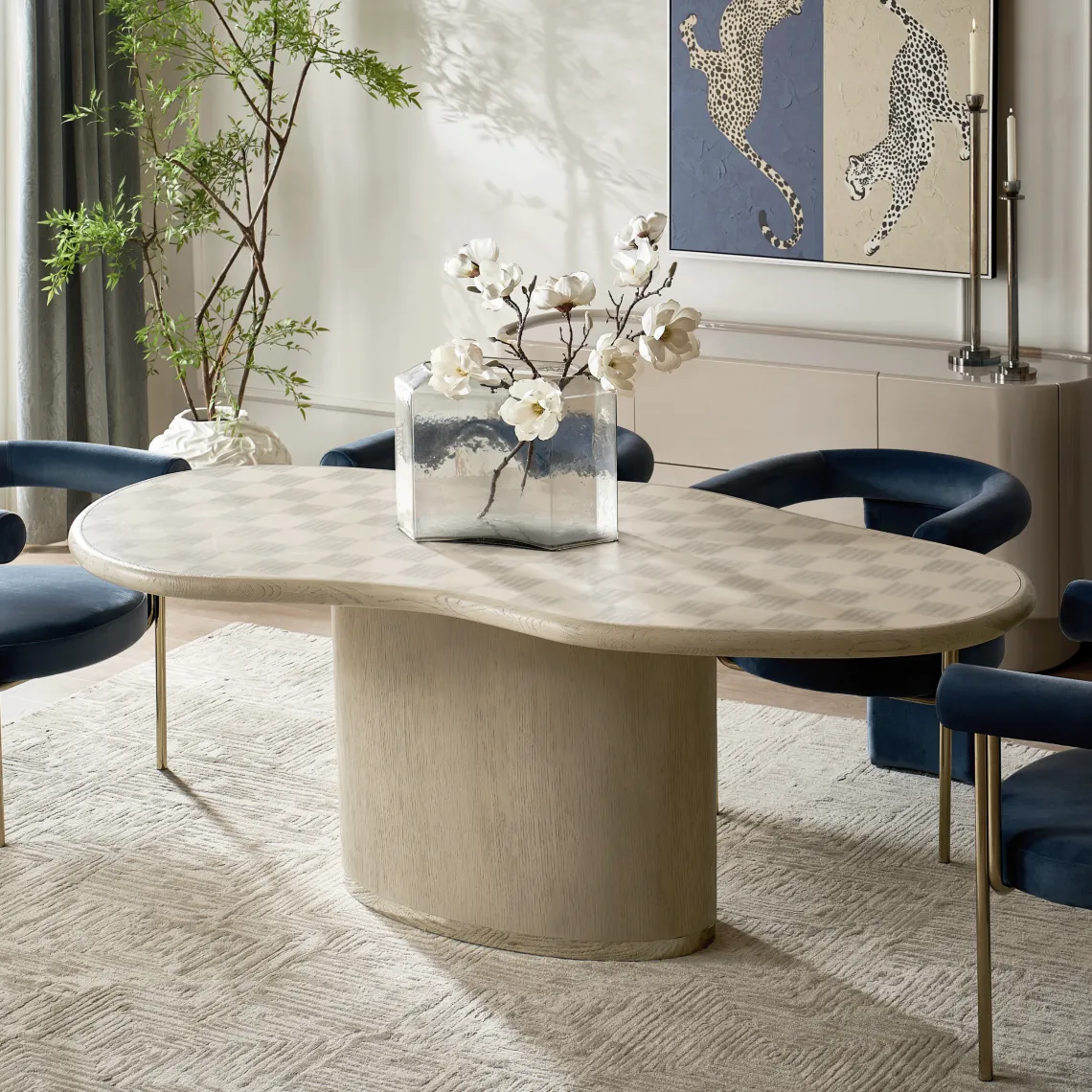 An organic kidney-shaped light oak dining table with a checkerboard parquet top, surrounded by navy blue velvet chairs in a bright modern dining room with a leopard art painting.