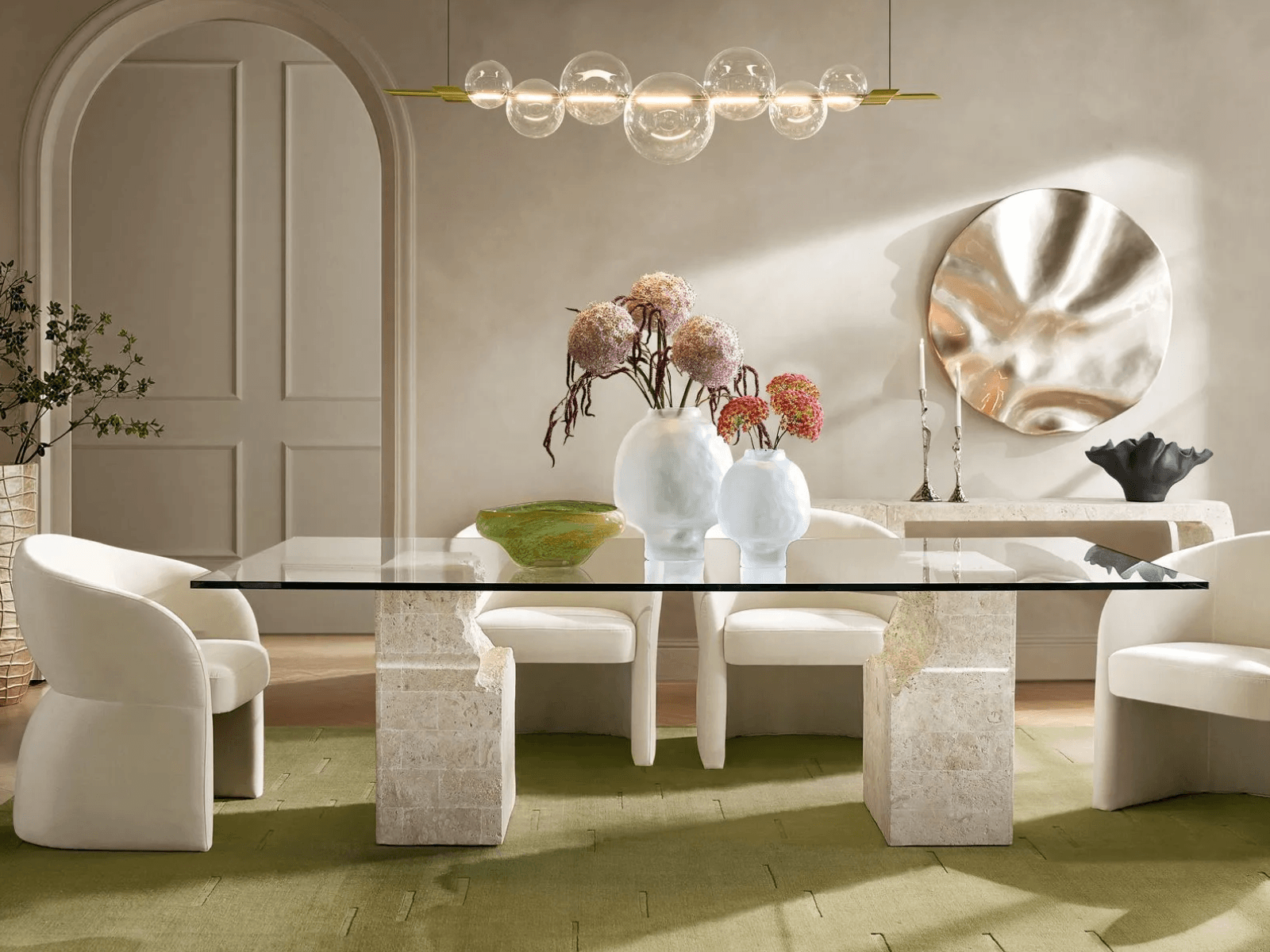 Contemporary dining space showcasing a clear glass top dining table with travertine stone pillars, curved white chairs, and a horizontal clear glass orb linear chandelier.