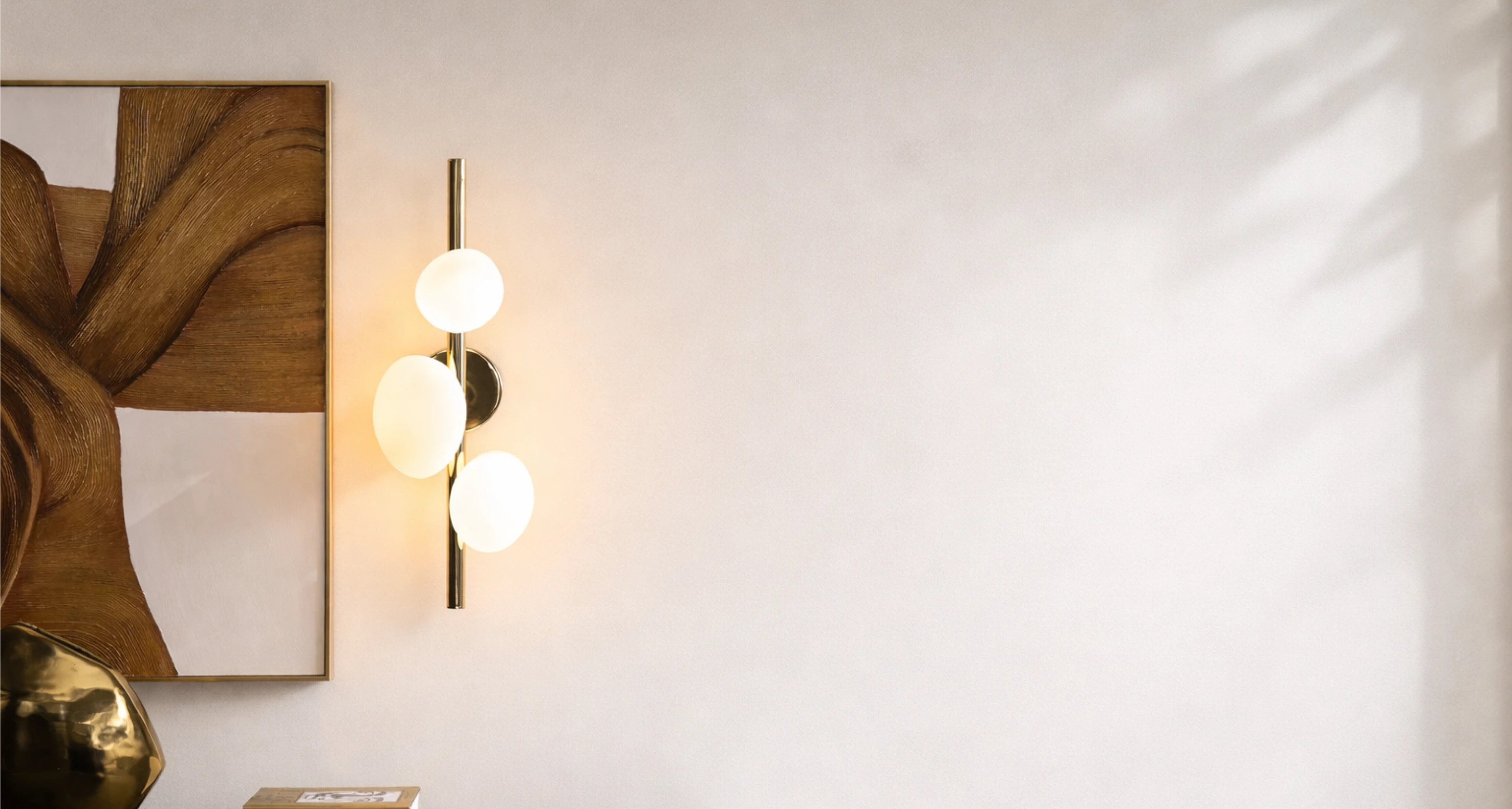 Modern vertical brass bar wall sconce featuring three illuminated frosted white orb globes mounted next to framed abstract canvas art.