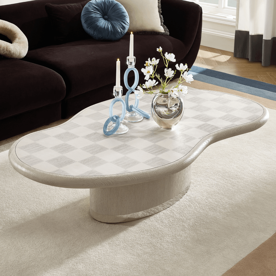 Sculptural curved organic coffee table with a subtle checkered top styled with blue loop candleholders and silver vase in front of a dark velvet sofa.