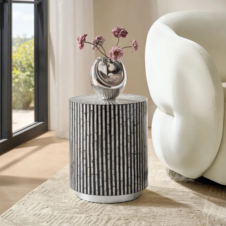 A cylindrical side table with black and white shell-inlay vertical stripes, topped with a fluid silver vase holding pink flowers, set beside a cream sofa.