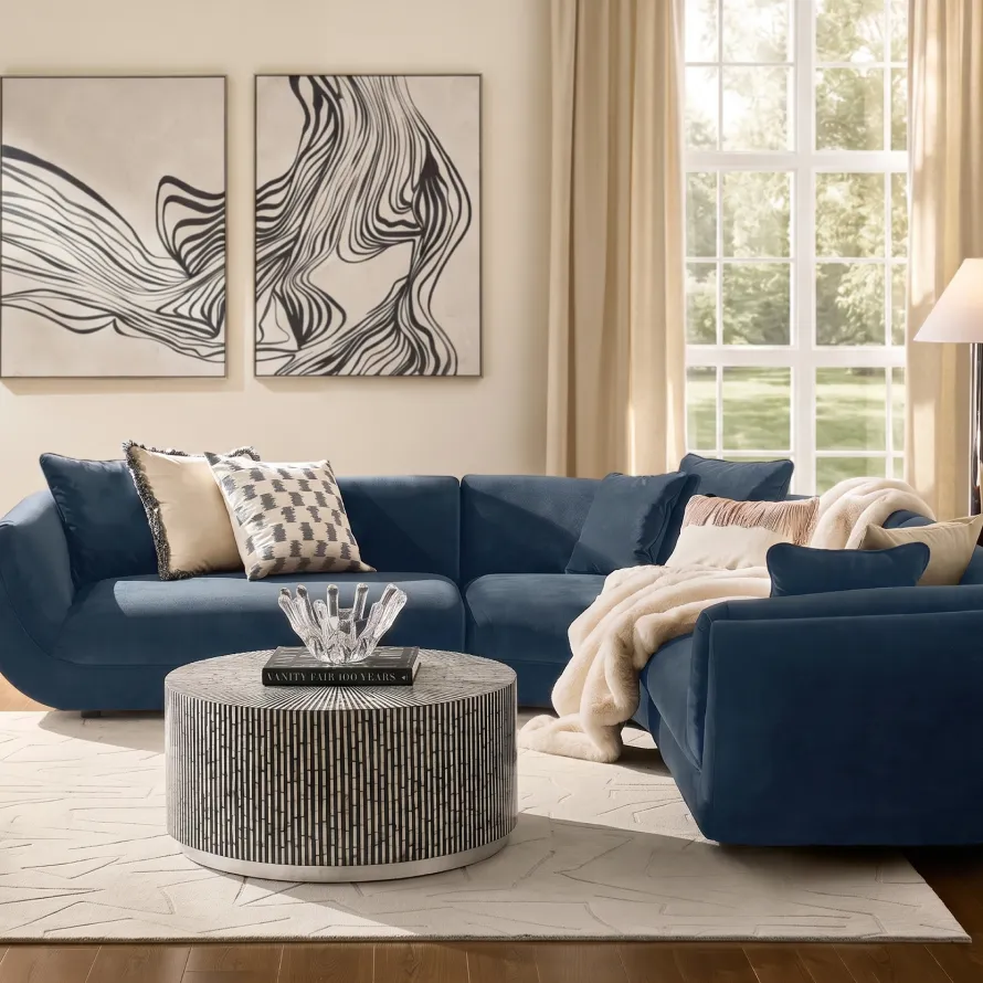 A modern luxury living room featuring a deep navy blue velvet sectional sofa, a black-and-white striped drum coffee table, diptych abstract wall art, and large sunlit windows.