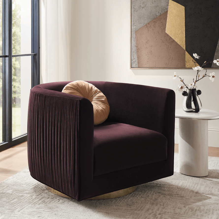 Deep burgundy velvet pleated barrel swivel chair with brushed brass base styled with a round accent pillow next to a modern side table.