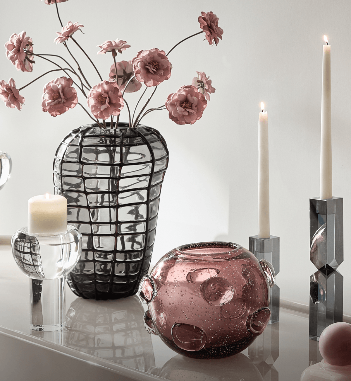 Elegant tabletop display featuring a dark grid-patterned glass vase with pink blooms, bubbly mauve glass bowl, crystal candle holders, and taper candles on a reflective console.