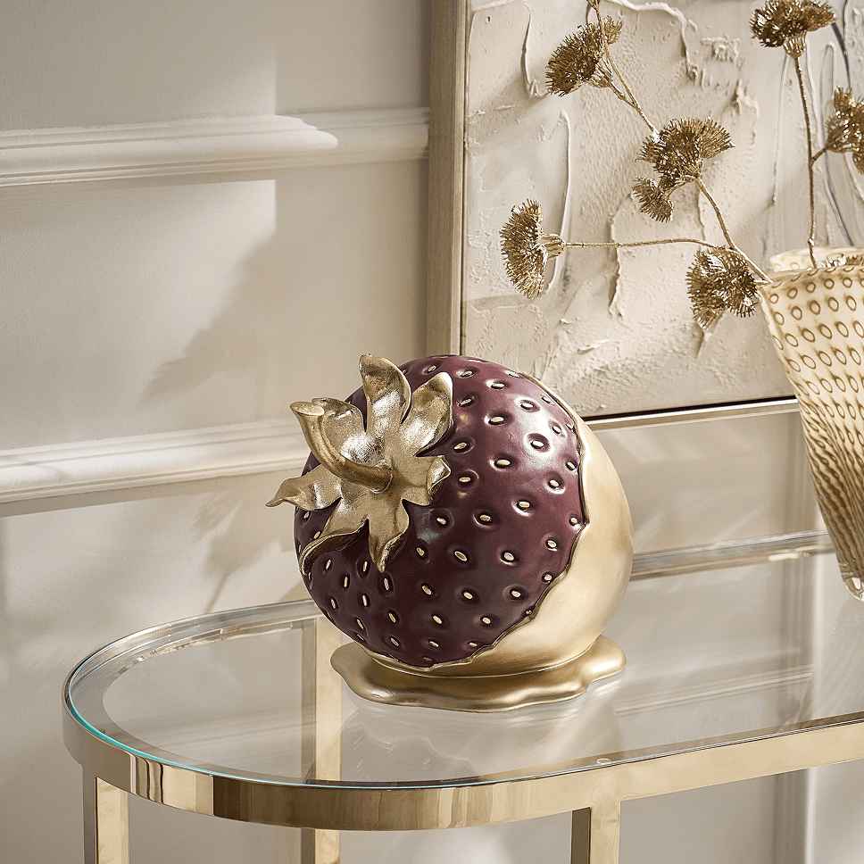 Sculptural metallic and deep plum strawberry decorative figurine displayed on a glass console table.