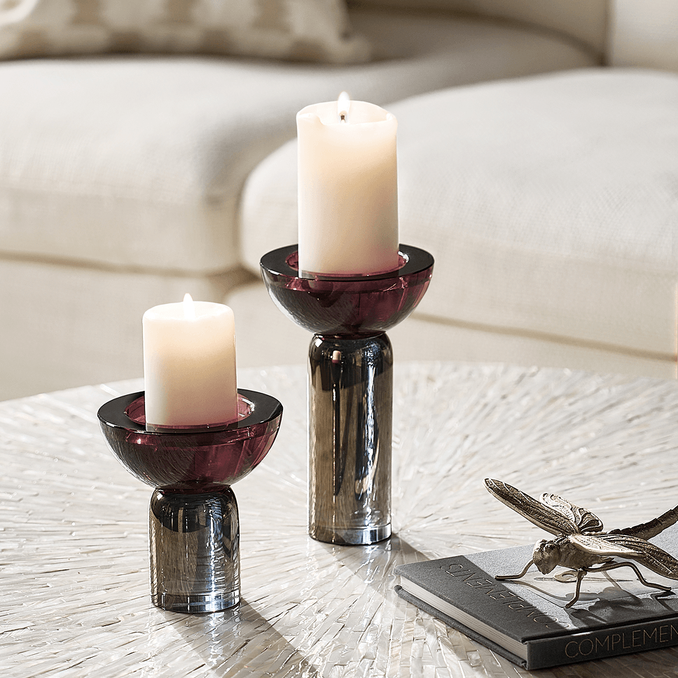 Pair of dark plum glass pillar candleholders with glowing white candles next to a metallic insect accent on a textured living room table.