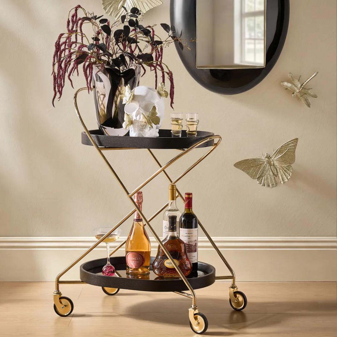 A luxury gold brass X-frame mobile bar cart with black trays, styled with liquor bottles, a floral vase, a butterfly-decorated skull, and wall-mounted gold metal butterflies.