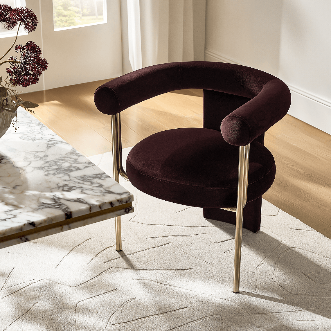 Deep burgundy velvet curved barrel dining chair with sleek brass legs positioned on an ivory geometric rug beside a white marble tabletop.