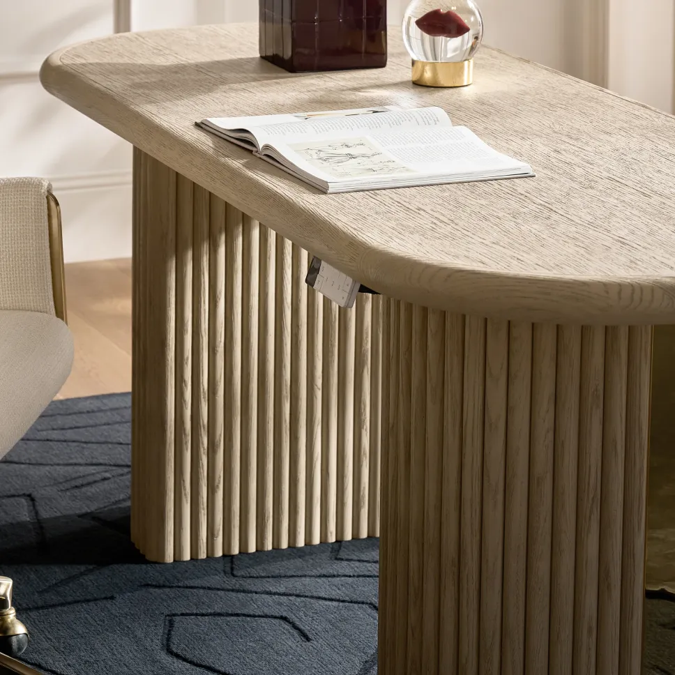 A close-up of a light oak executive desk with curved oval corners and fluted double-pedestal legs, topped with an open art book, a deep wine lamp base, and a glass lip paperweight.
