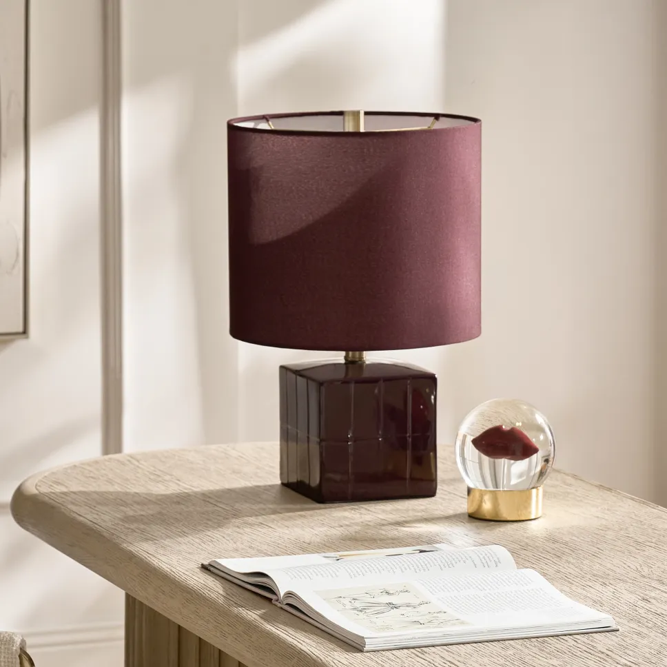 A deep burgundy table lamp with a matching shade and glossy cube base sitting on a light oak desk next to a red lips glass globe paperweight.