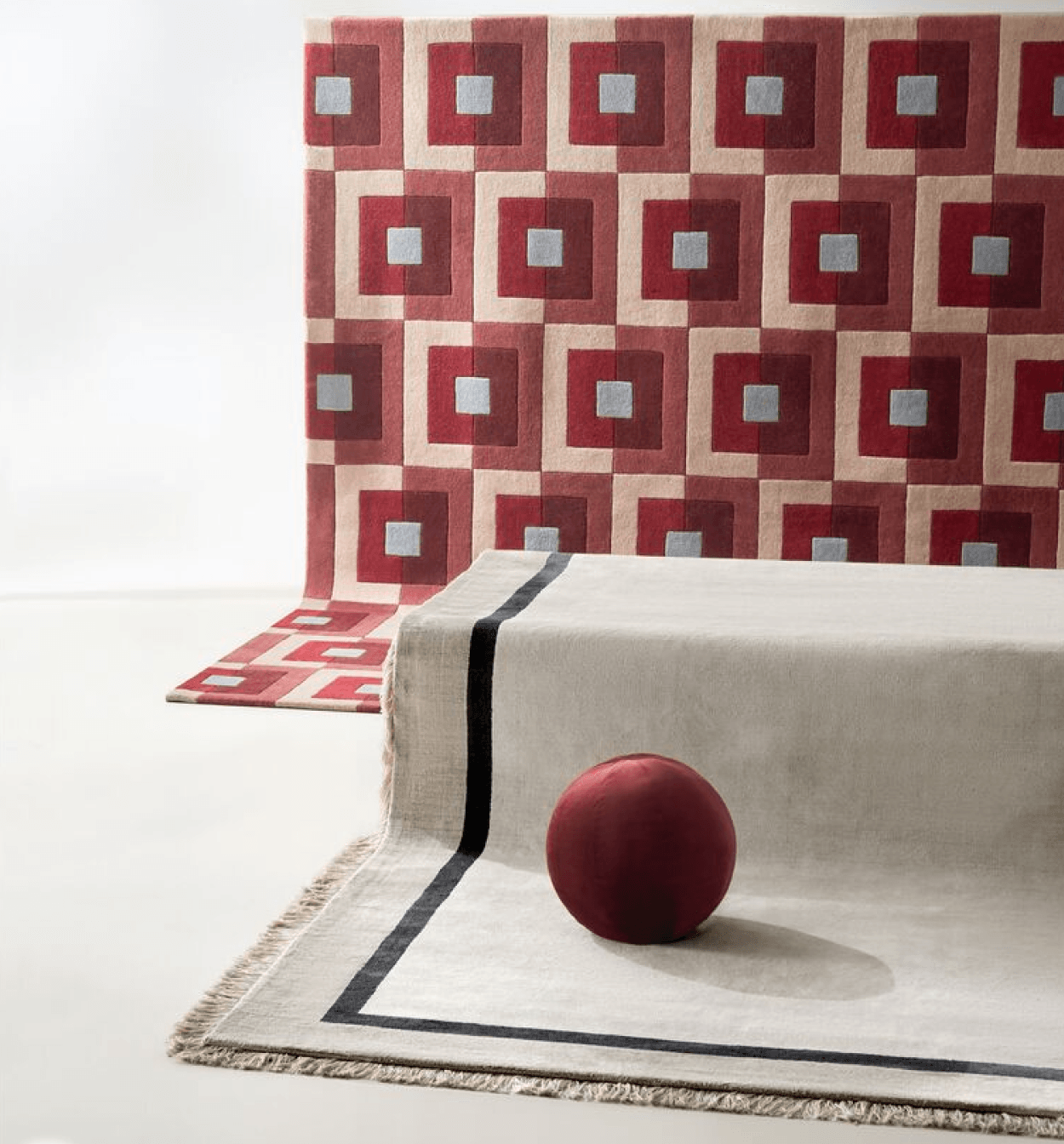 Contemporary geometric wool area rugs featuring a red and blush pink square optical pattern rug draped behind a light grey fringed rug accented with a black border and burgundy sphere.