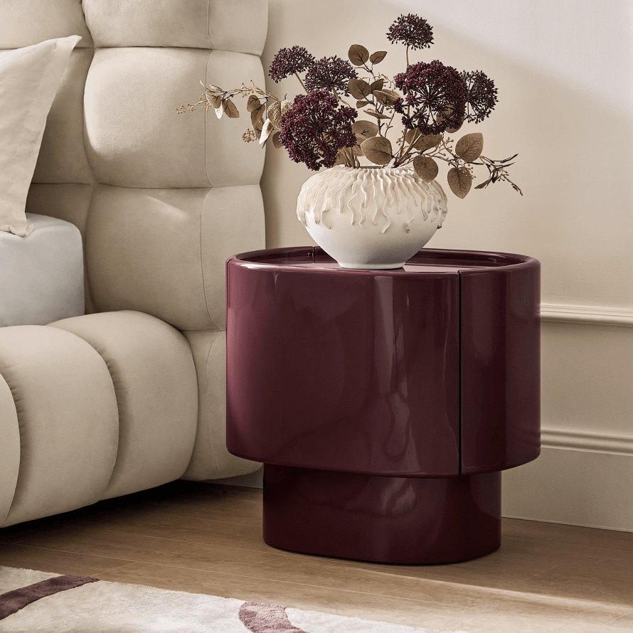 Deep burgundy high-gloss lacquered curved nightstand topped with floral decor in a textured white vase beside a channeled upholstered bed.