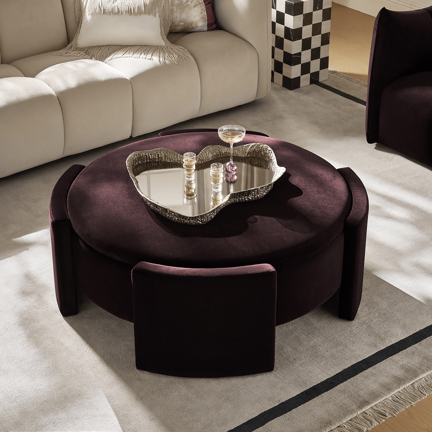 Modern living room featuring a plush deep plum round ottoman coffee table with nesting stools and styled drink tray, paired with a cream sofa and checkered side table.