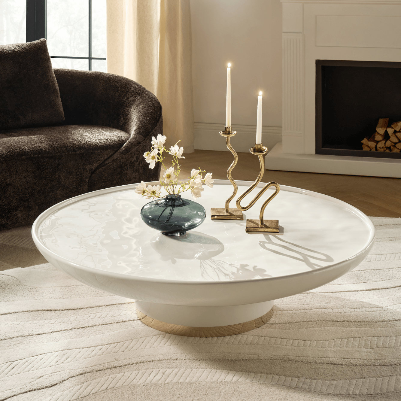 Large round off-white pedestal coffee table styled with brass candle holders, a small glass vase, and a dark plush armchair in a living room.
