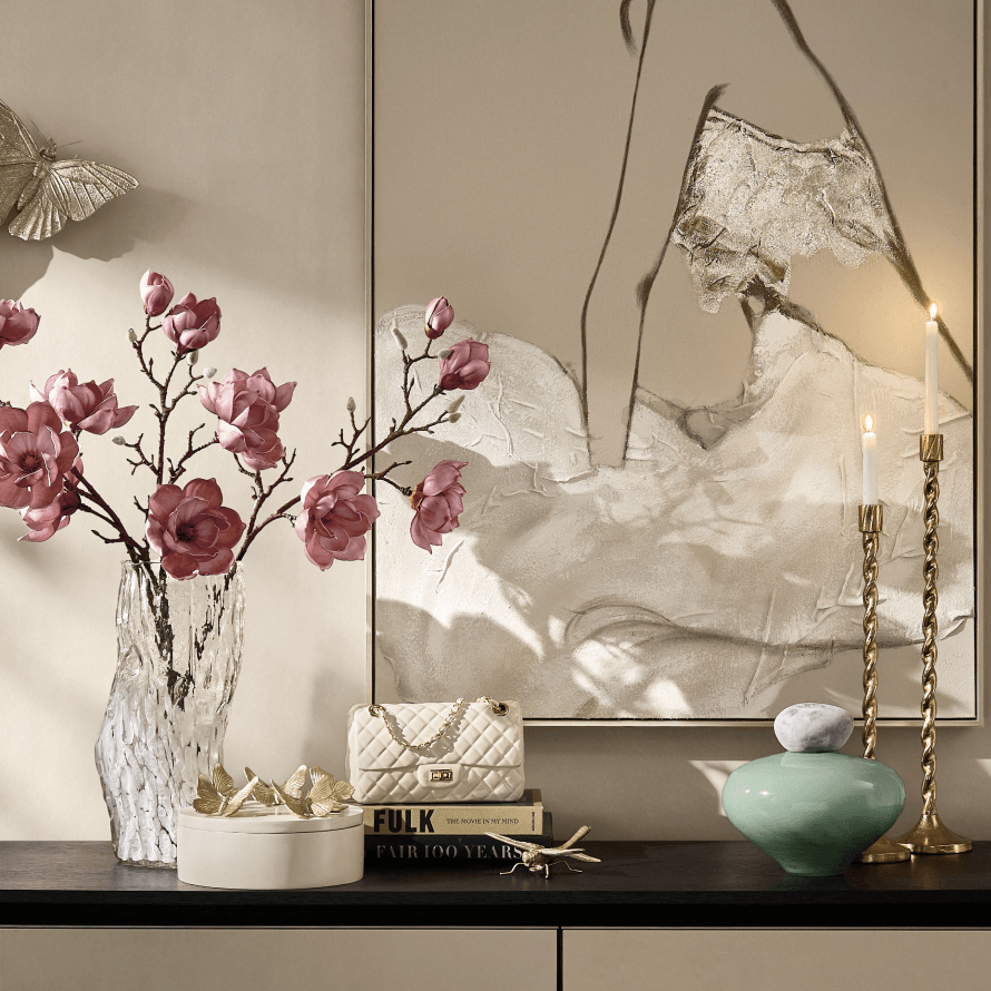 Elegant console tabletop styling featuring fresh pink blossoms in a crystal vase, neutral figurative wall art, brass candlesticks, and sculpted decorative accents.