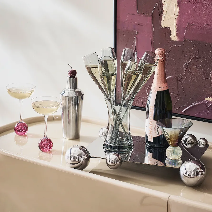 Luxury bar setup featuring glass coupe cocktail glasses with pink globe stems, polka-dot champagne flutes in a vase, a chrome shaker, and a bottle of Chandon sparkling wine on a mirrored tray.