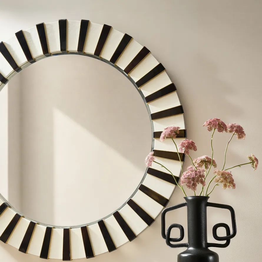 A close-up of a round Art Deco mirror with a black and cream striped sunburst frame, next to a matte black sculptural vase with pink flowers.