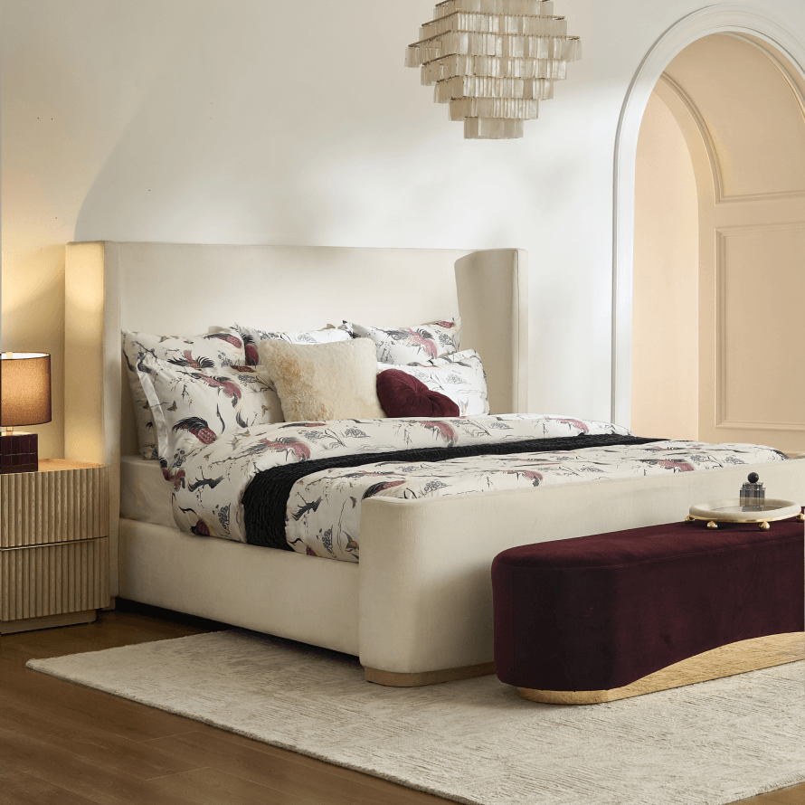 Modern upholstered cream shelter bed styled with patterned bedding, a tiered crystal chandelier, and a deep burgundy velvet end-of-bed bench.