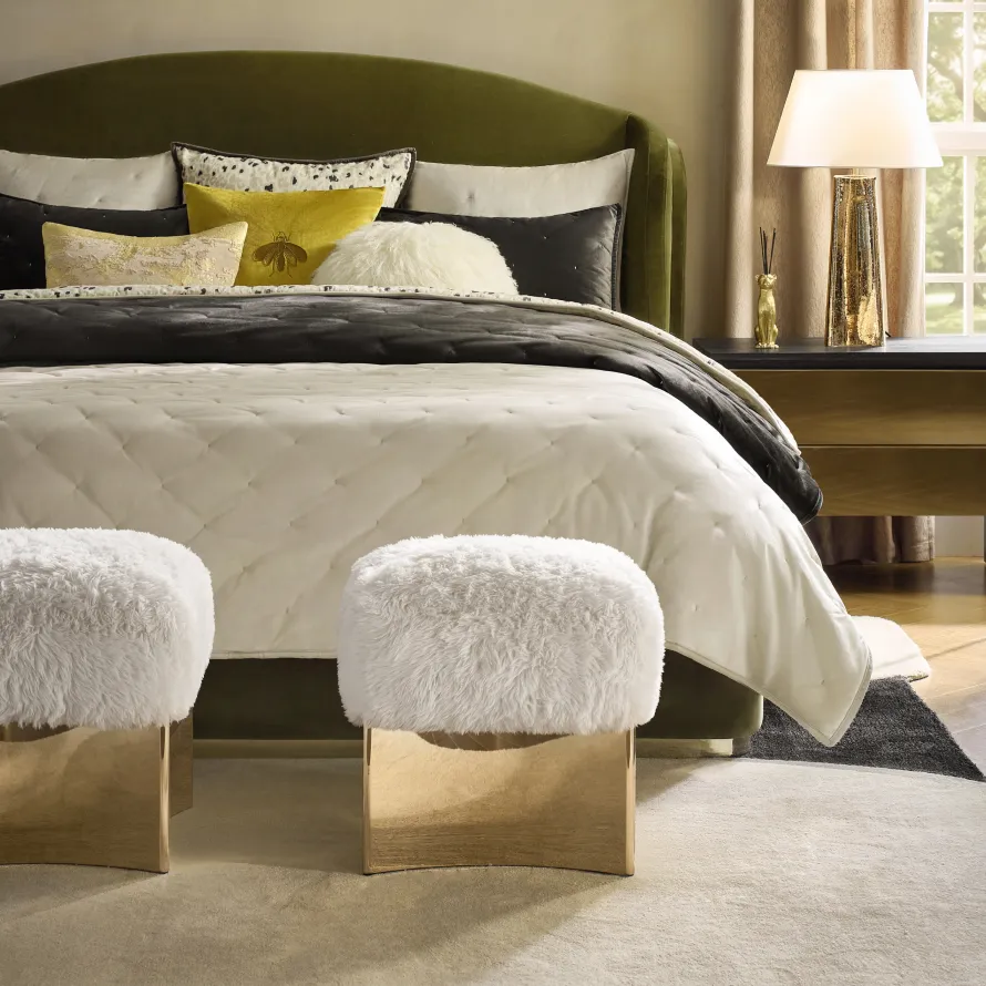 A luxury bedroom scene featuring a curved olive green velvet bed, white faux-fur ottomans with brass bases, quilted cream bedding, and a gold table lamp.