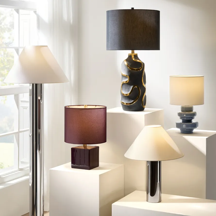 A curated display of modern designer lamps, including a chrome floor lamp, an organic black ceramic lamp with gold accents, and a burgundy block table lamp on white plinths.