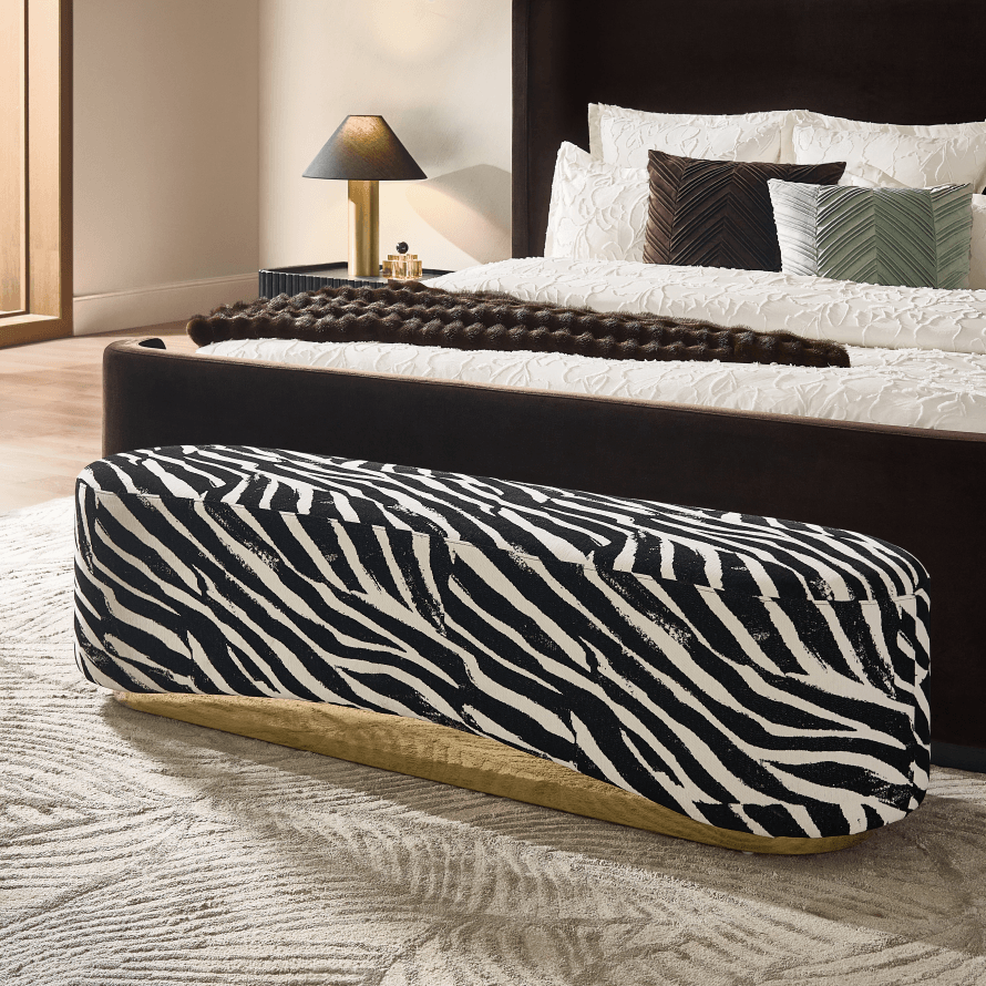 Modern bedroom bench in bold black and white zebra print with a brass base set at the foot of a dark upholstered bed on a textured rug.