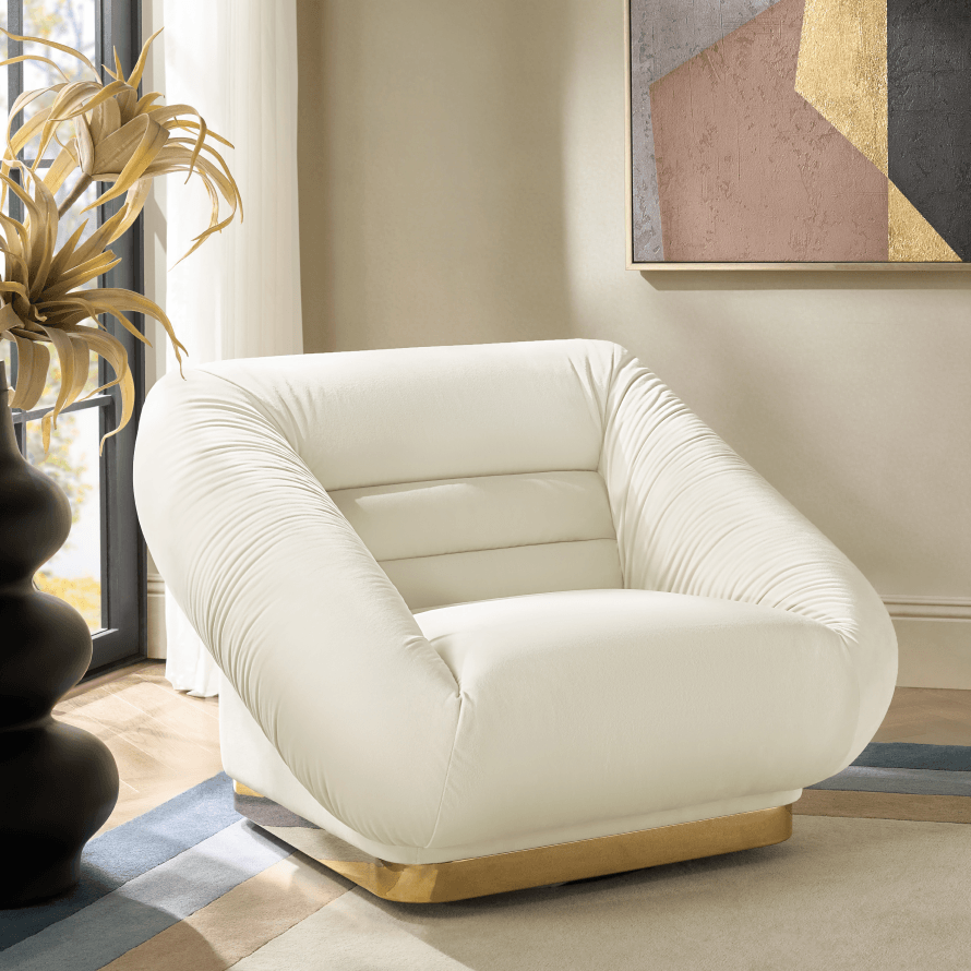 Modern off-white channel-tufted accent chair with plissé armrests and a brass pedestal base, styled next to a black floor vase and abstract wall art.