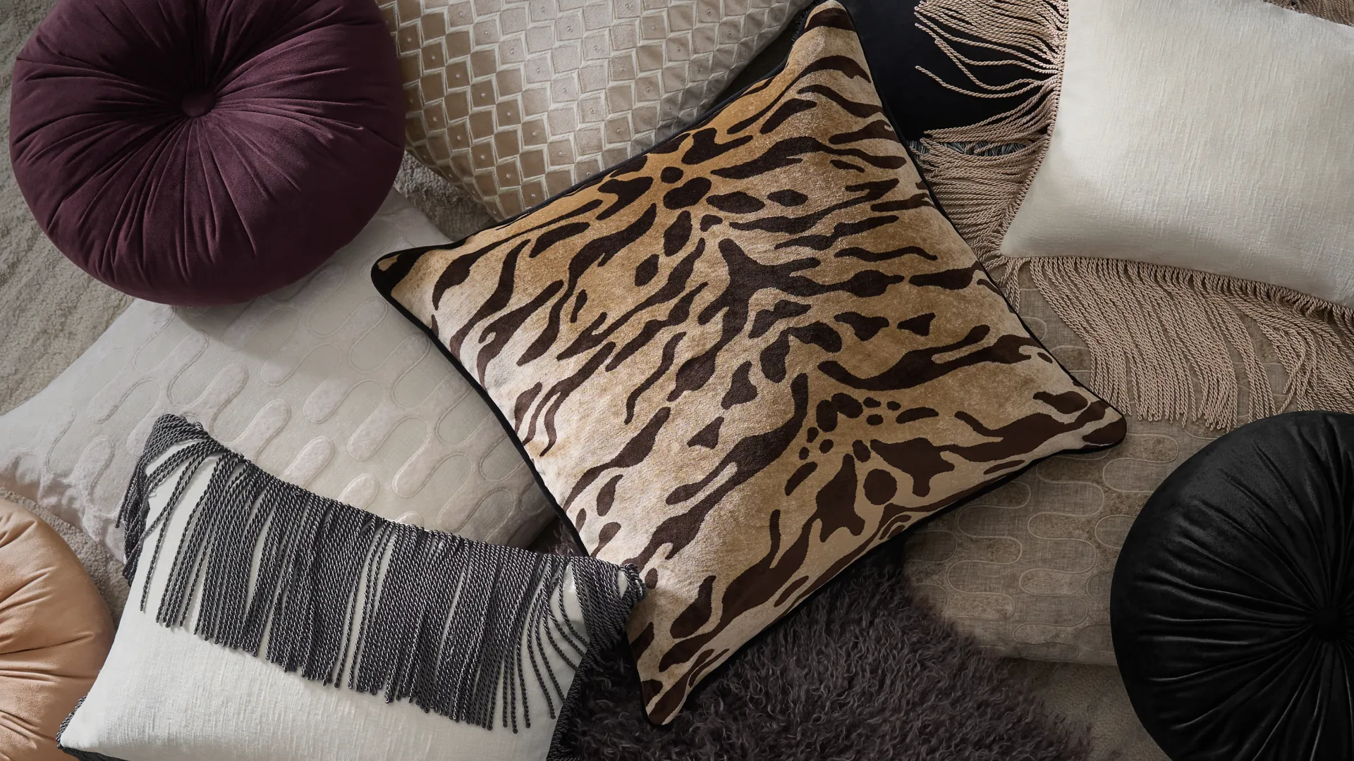 A luxury flatlay of textured throw pillows featuring a tiger-striped cushion surrounded by deep plum round velvet pillows, fringed cushions, and faux-fur textiles.
