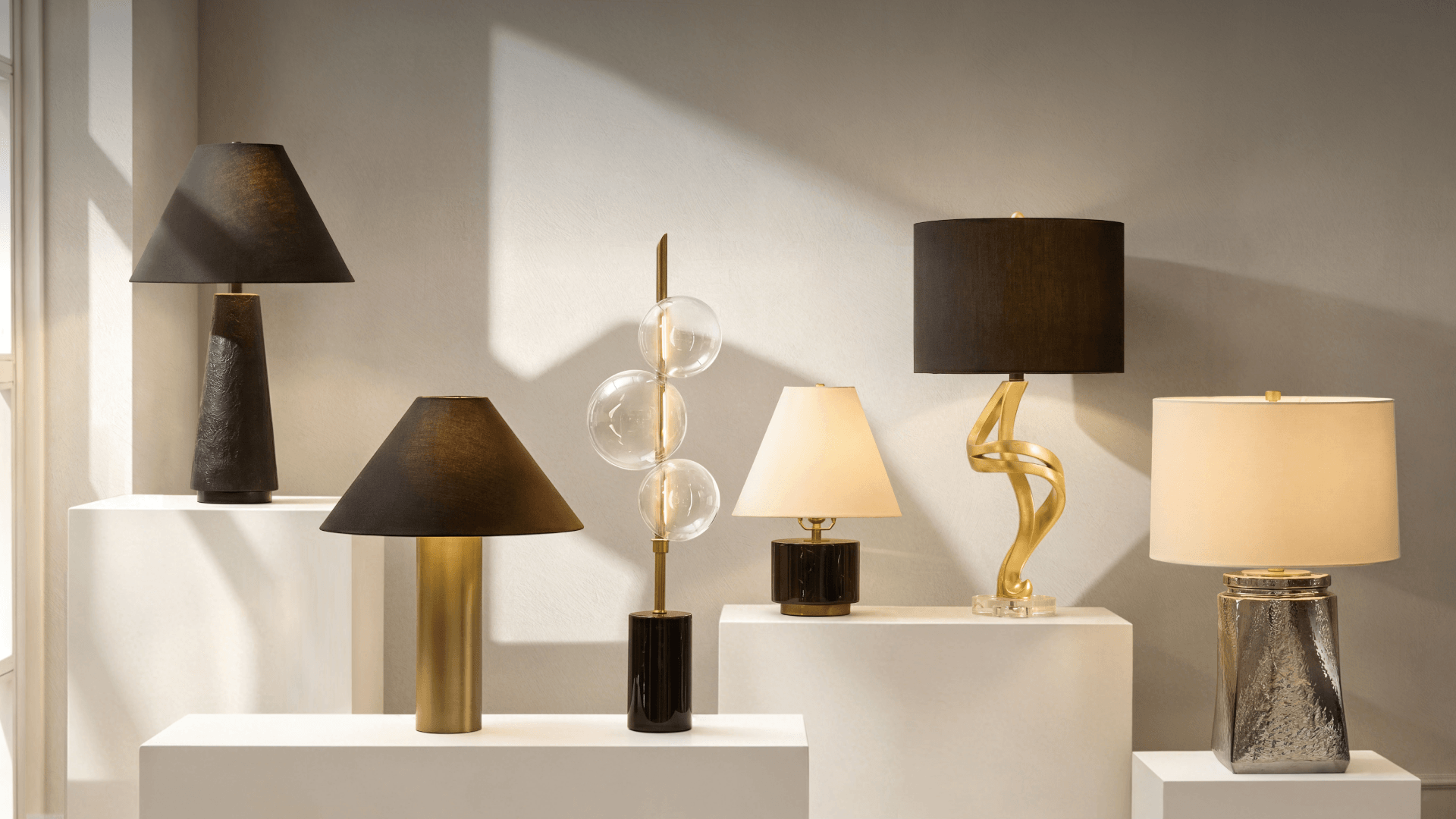 Collection of luxury table lamps featuring sculpted brass bases, black shades, glass globe accents, and polished ceramic finishes displayed on white pedestals.
