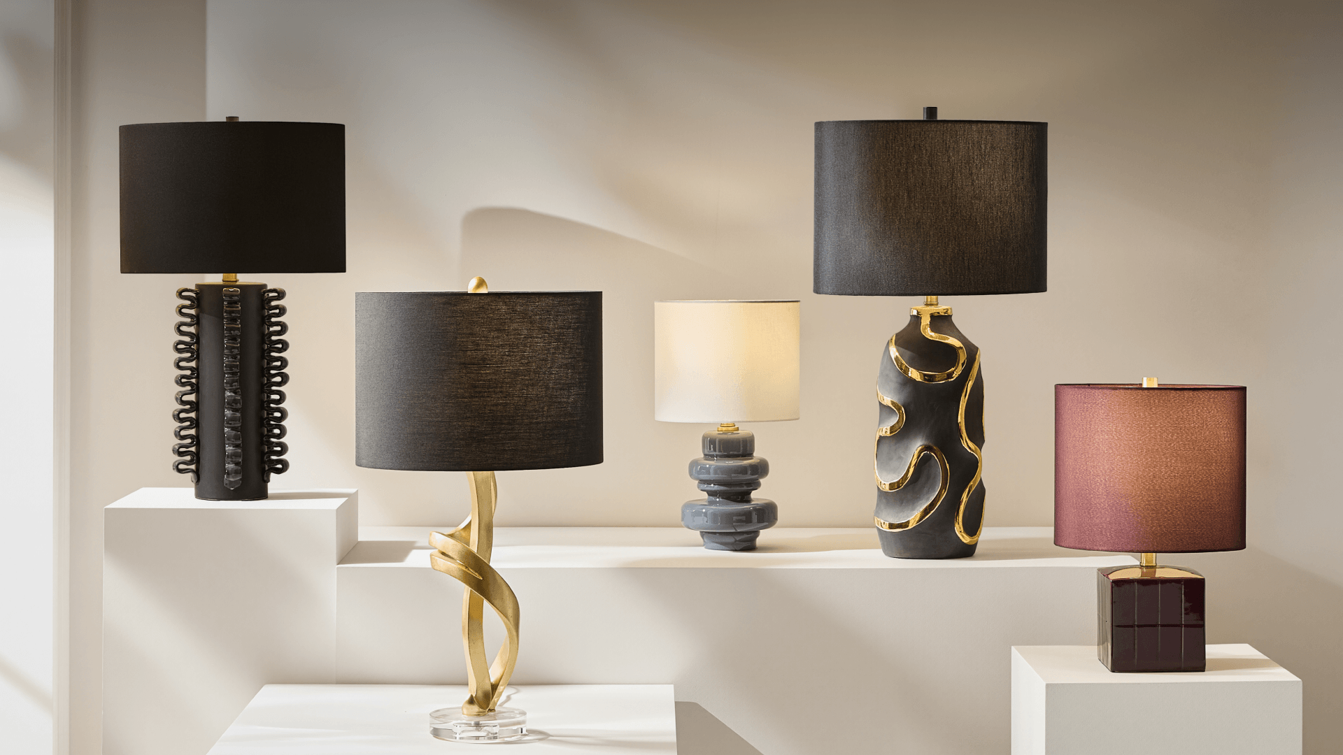 Designer table lamp display featuring sculpted modern bases in matte black, twisted gold, stacked gray ceramic, and deep plum gloss finishes on white gallery pedestals.
