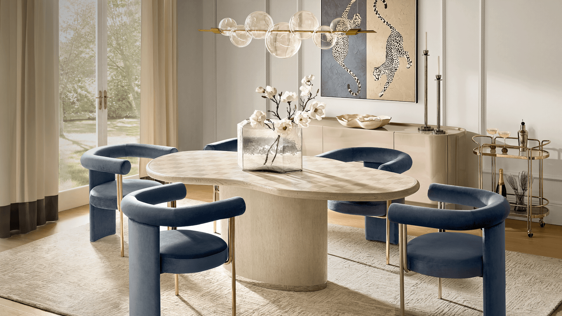 Modern dining room featuring a light wood pedestal table with subtle checkered top, blue velvet curved dining chairs, a linear bubble glass chandelier, and a rolling brass bar cart in front of cheetah artwork.