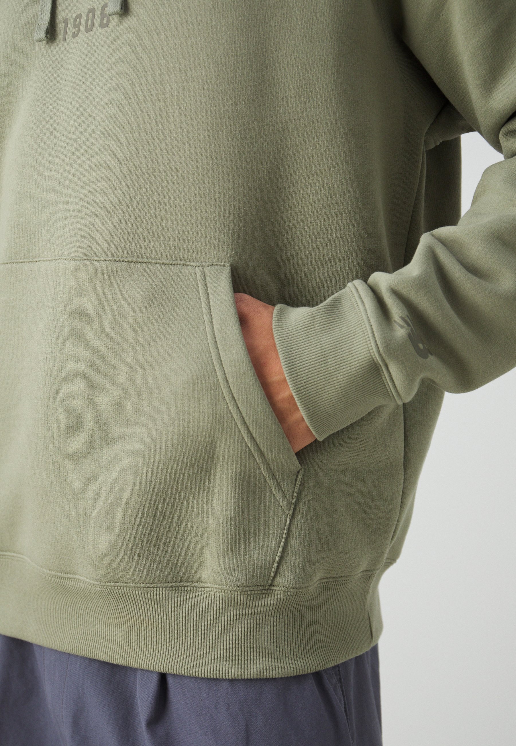Sudadera verde caqui hecha de una tela suave, con un bolsillo frontal y puños acanalados. Presenta un diseño minimalista y un pequeño texto en relieve.