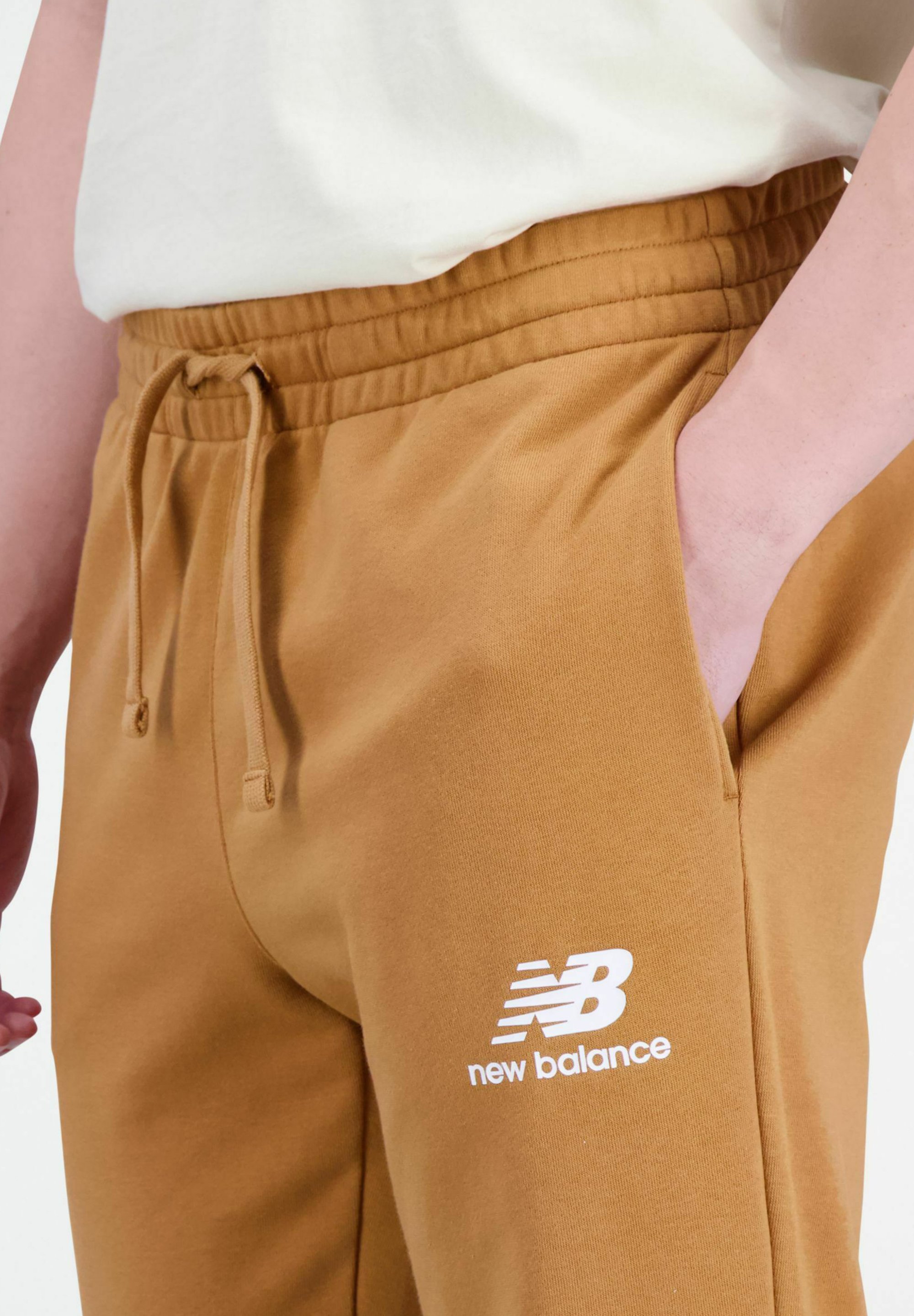 Pantalones jogger marrones hechos de material suave con una cinturilla elástica, que cuentan con un cordón y un logo blanco de "New Balance" en el muslo.