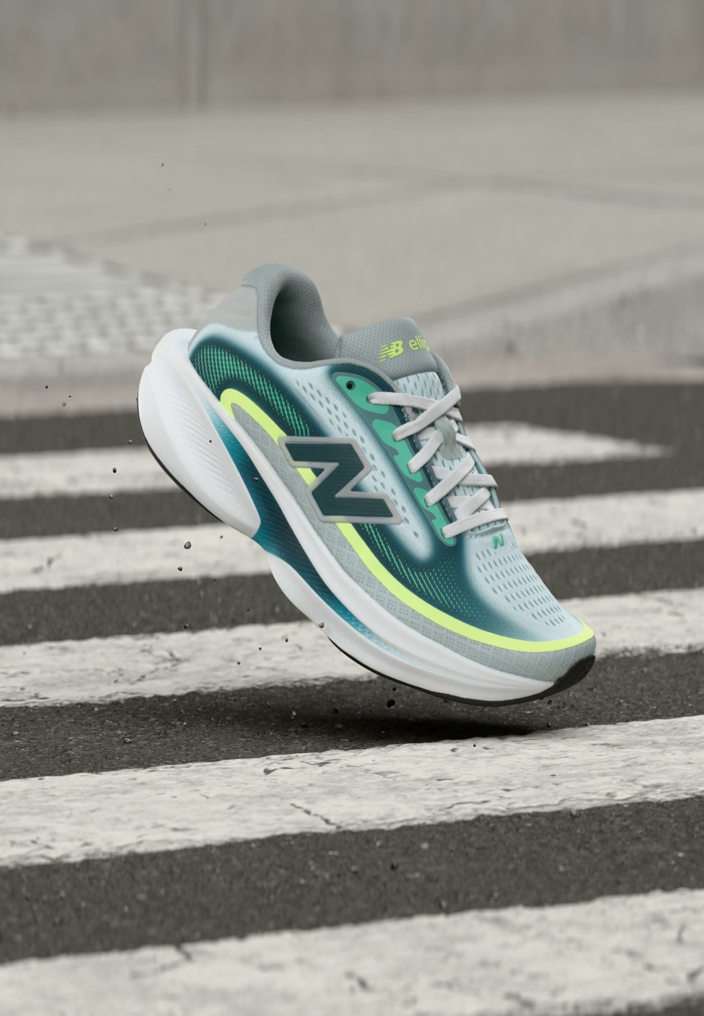 Zapatilla de running New Balance en azul claro y verde con suela blanca, mostrada en el aire sobre un paso de peatones de cebra en un pavimento de asfalto.