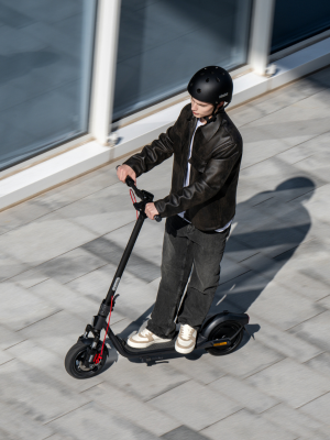 Persona in sella a uno scooter elettrico su una superficie asfaltata accanto a finestre di vetro, con giacca scura e abbigliamento casual.