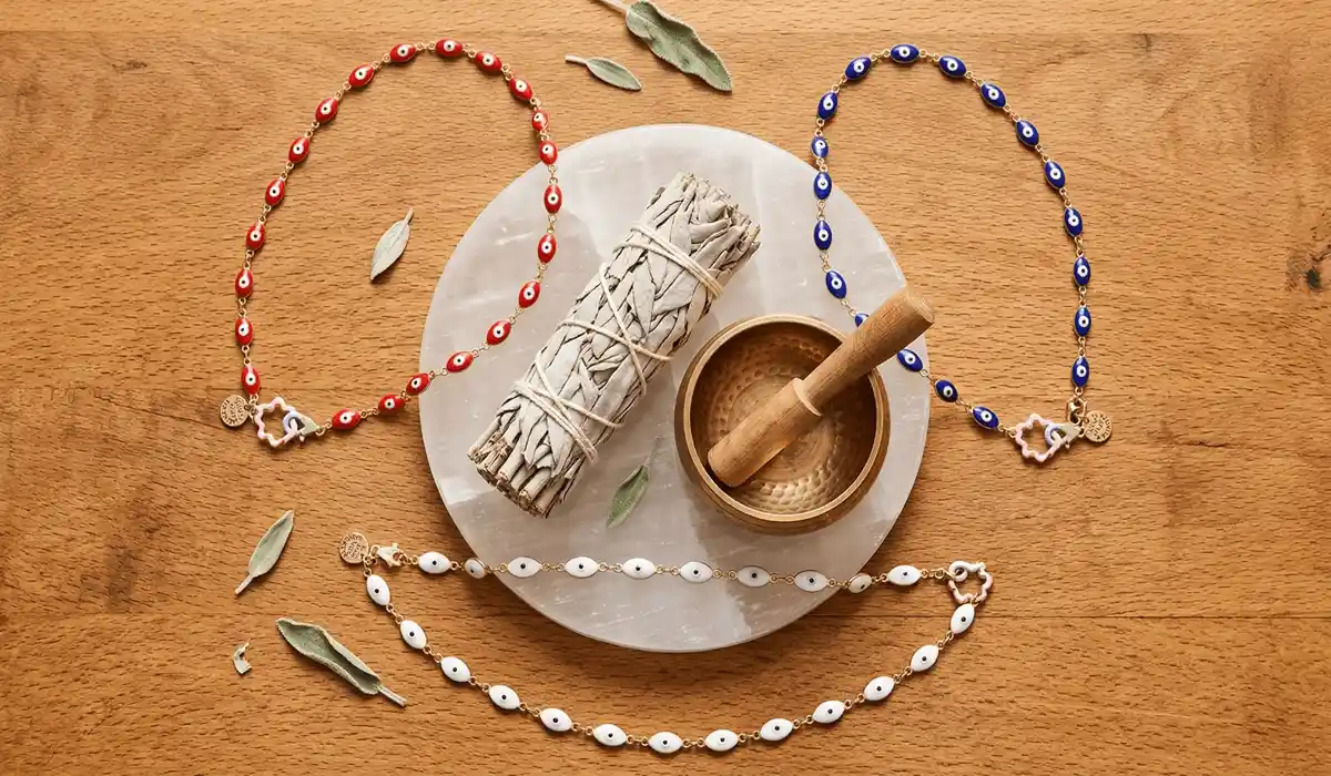 Different colors of evil eye necklaces including red, blue, and white, arranged with cleansing tools like sage and selenite.