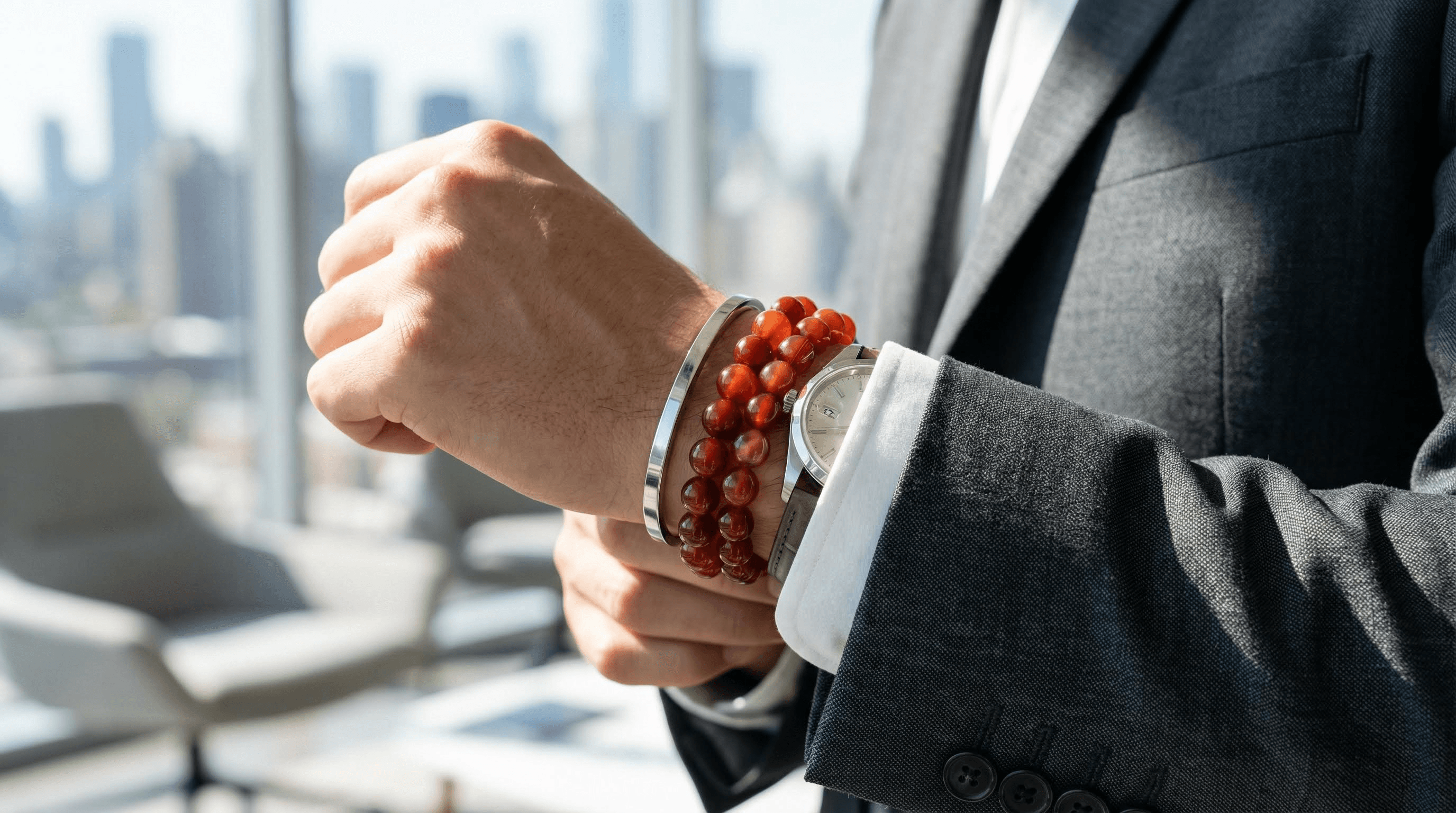A person's wrist wearing a Red Agate and silver bracelet stack with a luxury watch and a business suit.