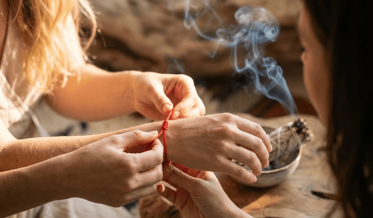 A spiritual cleansing ritual featuring a loved one tying a red string bracelet on a wrist with sage smoke in the background.