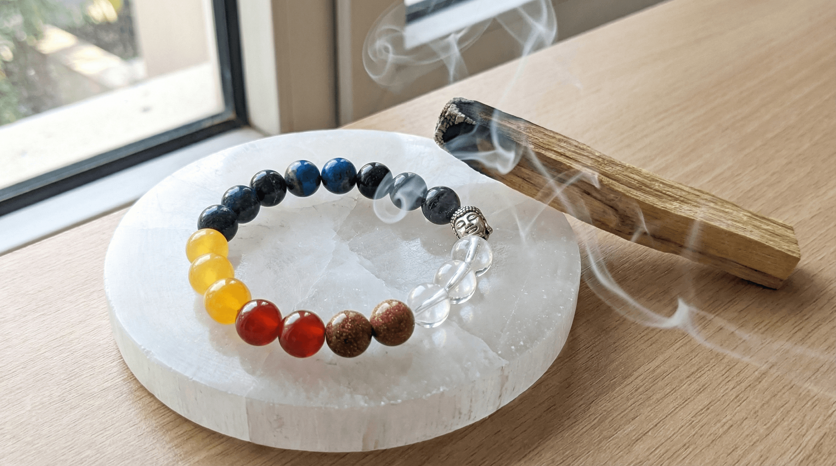 A 7 chakra bracelet being cleansed with Palo Santo smoke and charged on a selenite plate for spiritual activation.