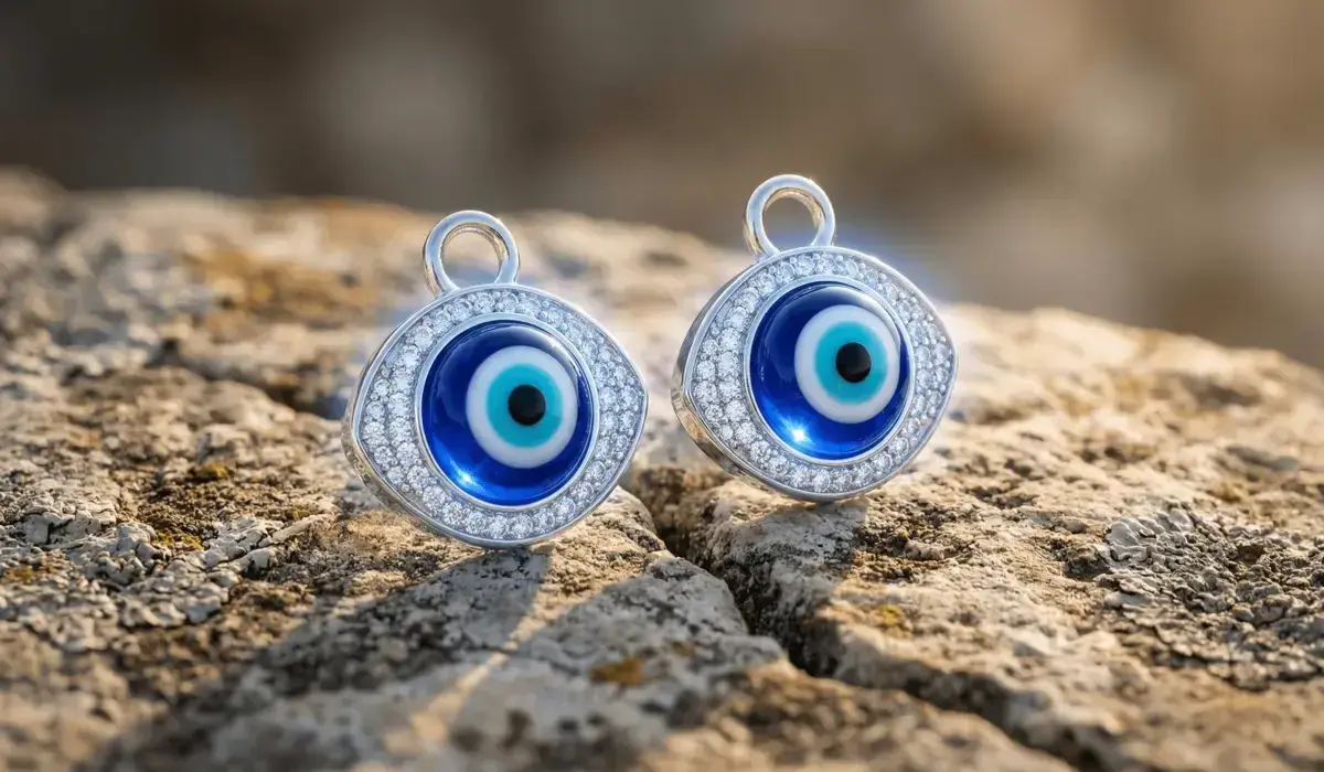 Pair of silver Evil Eye pendants encrusted with cubic zirconia, featuring classic blue and white evil eye centers, resting on a textured rocky surface under warm natural sunlight.
