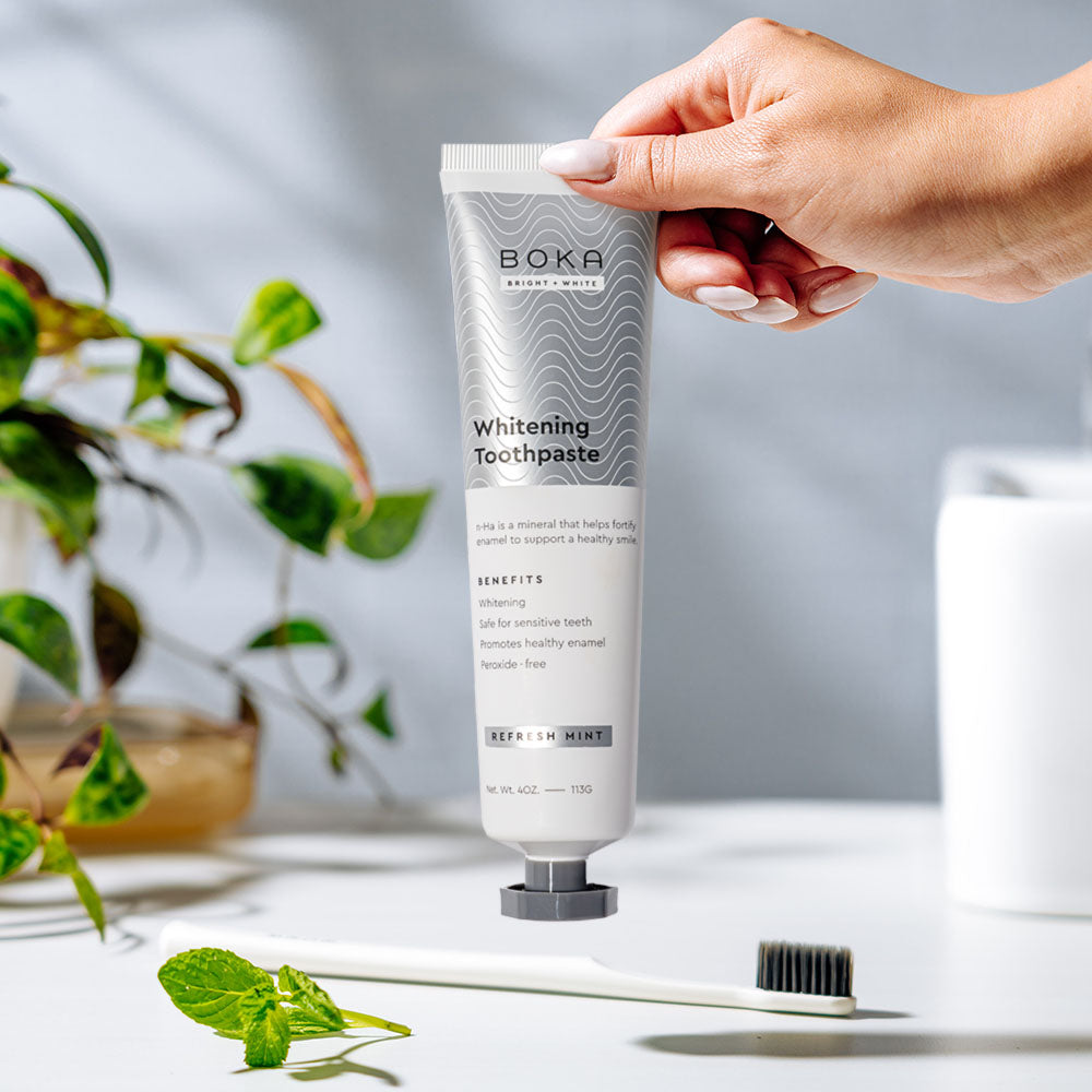 A hand holds Nano (n-Ha) Toothpaste Whitening 3 Pack in Refresh Mint above a toothbrush on white surface.
