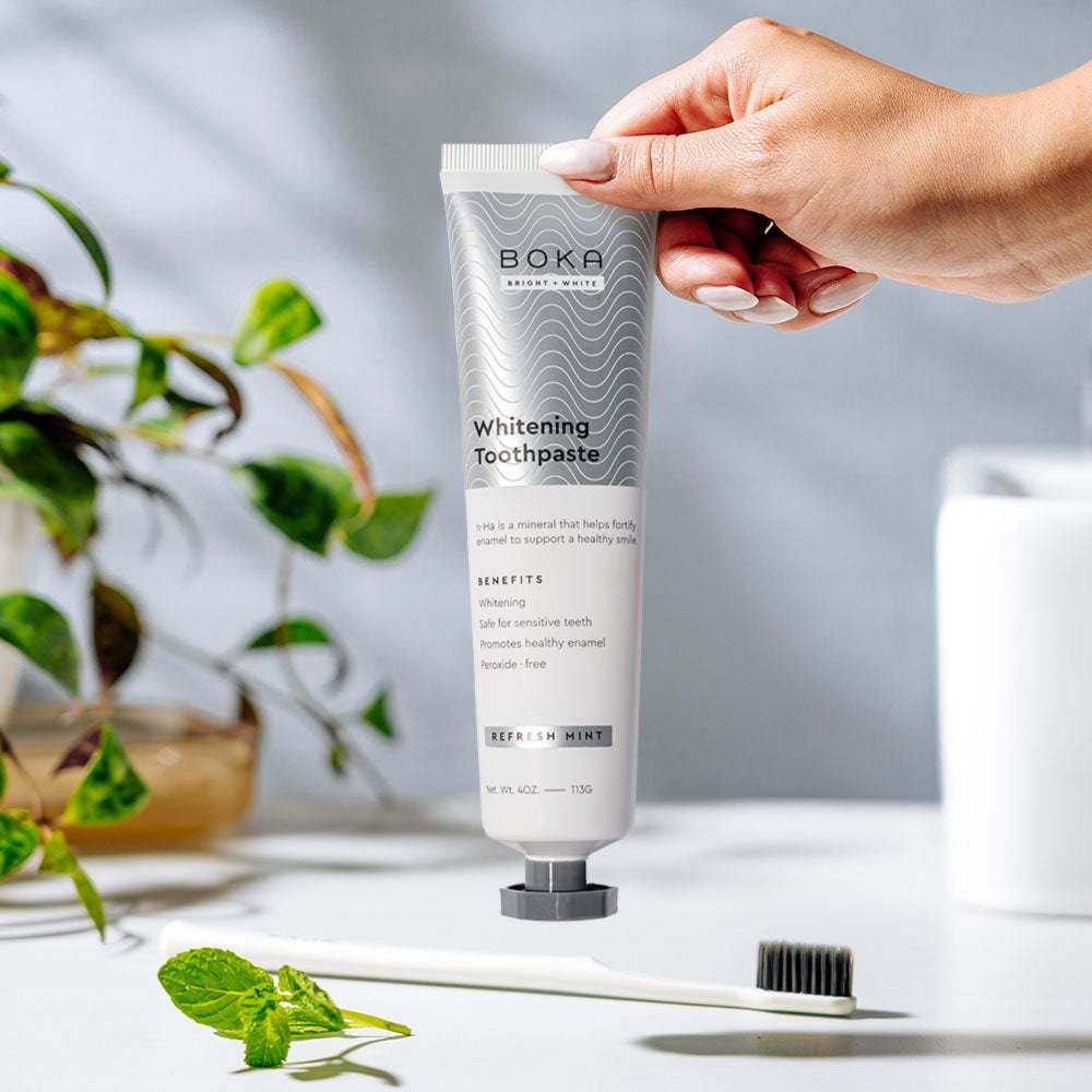 A hand holds Nano (n-Ha) Toothpaste Whitening 3 Pack in Refresh Mint above a toothbrush on white surface.