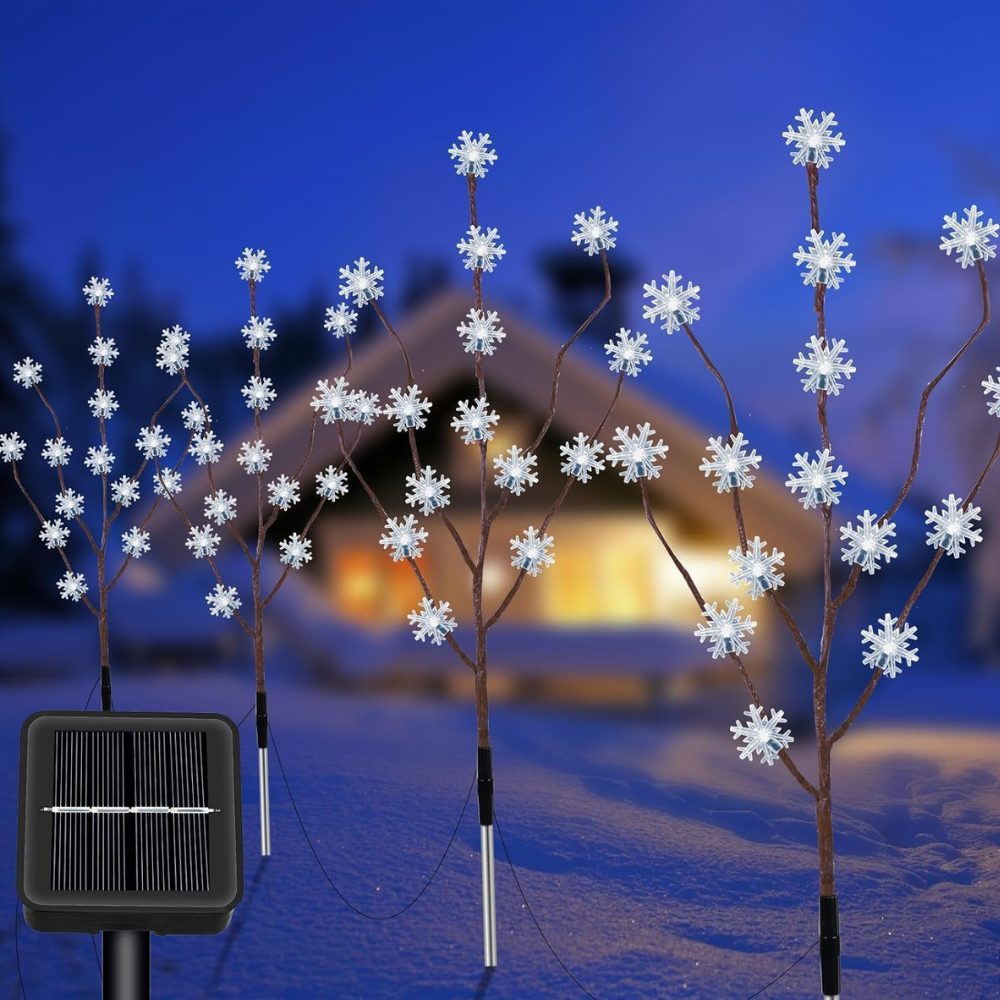 Two solar snowflake branch pathway lights with white snowflake-shaped LEDs staked in snow beside a winter chalet, solar panel visible