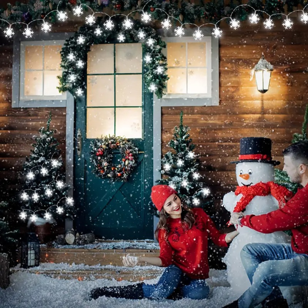 LED snowflake curtain string lights in cool white draped over a snowy cabin entrance with a couple and snowman