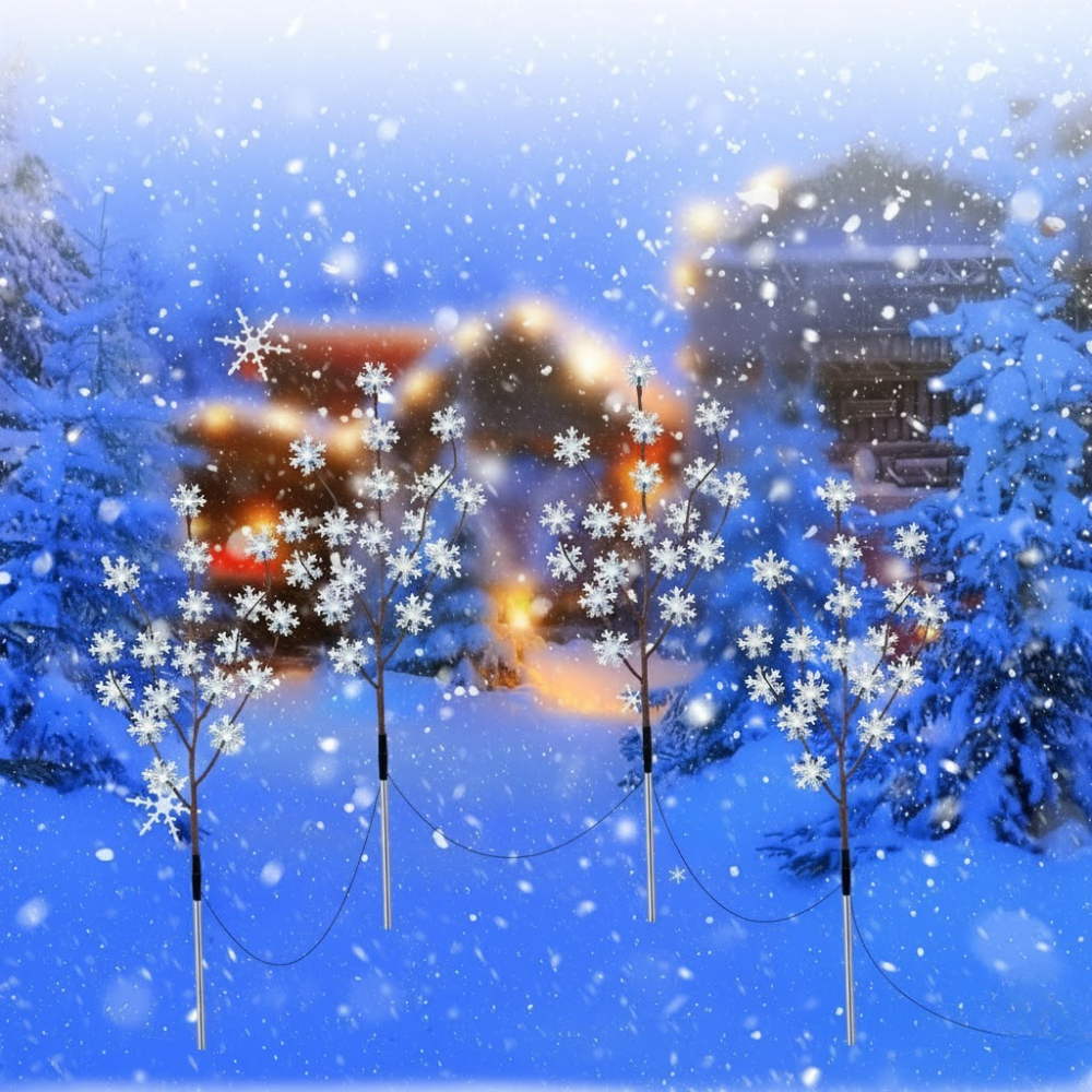 Three solar snowflake branch outdoor lights glowing white against a deep blue winter sky with snow and a distant lit cabin