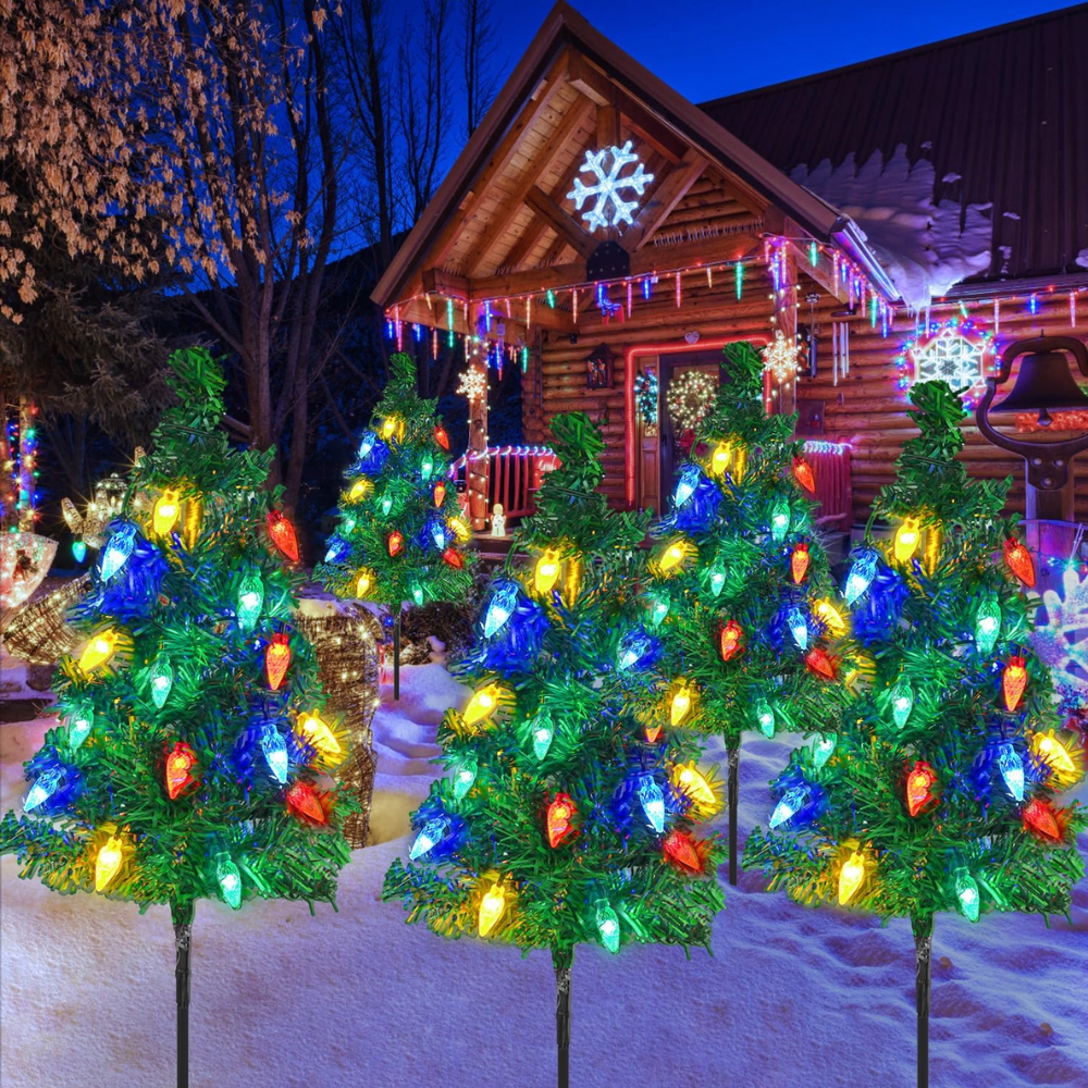 Three solar Christmas tree pathway lights with blue and multicolor LED ornaments staked in snow beside a log cabin with extensive holiday light displays