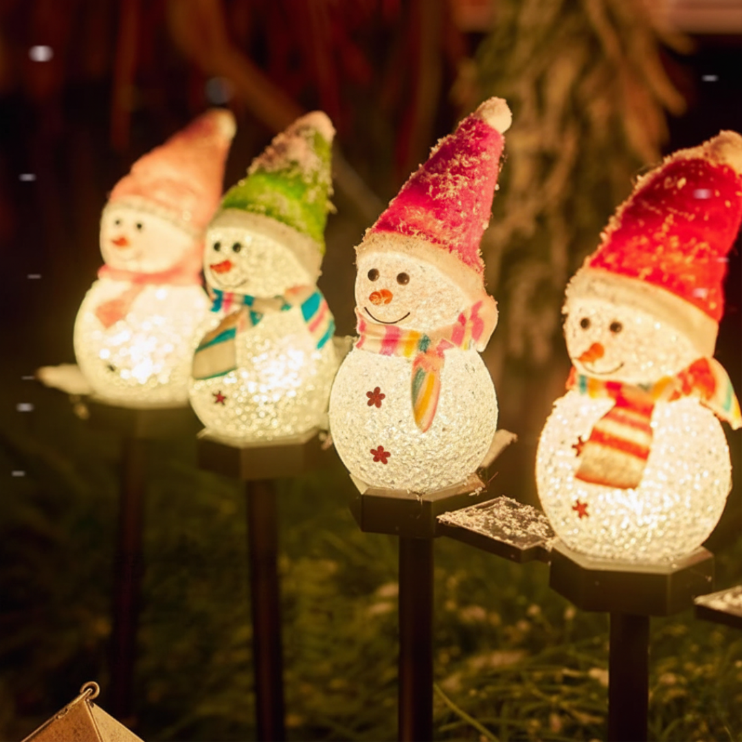 Four solar snowman Christmas pathway lights in pastel hats glowing at night in a garden setting