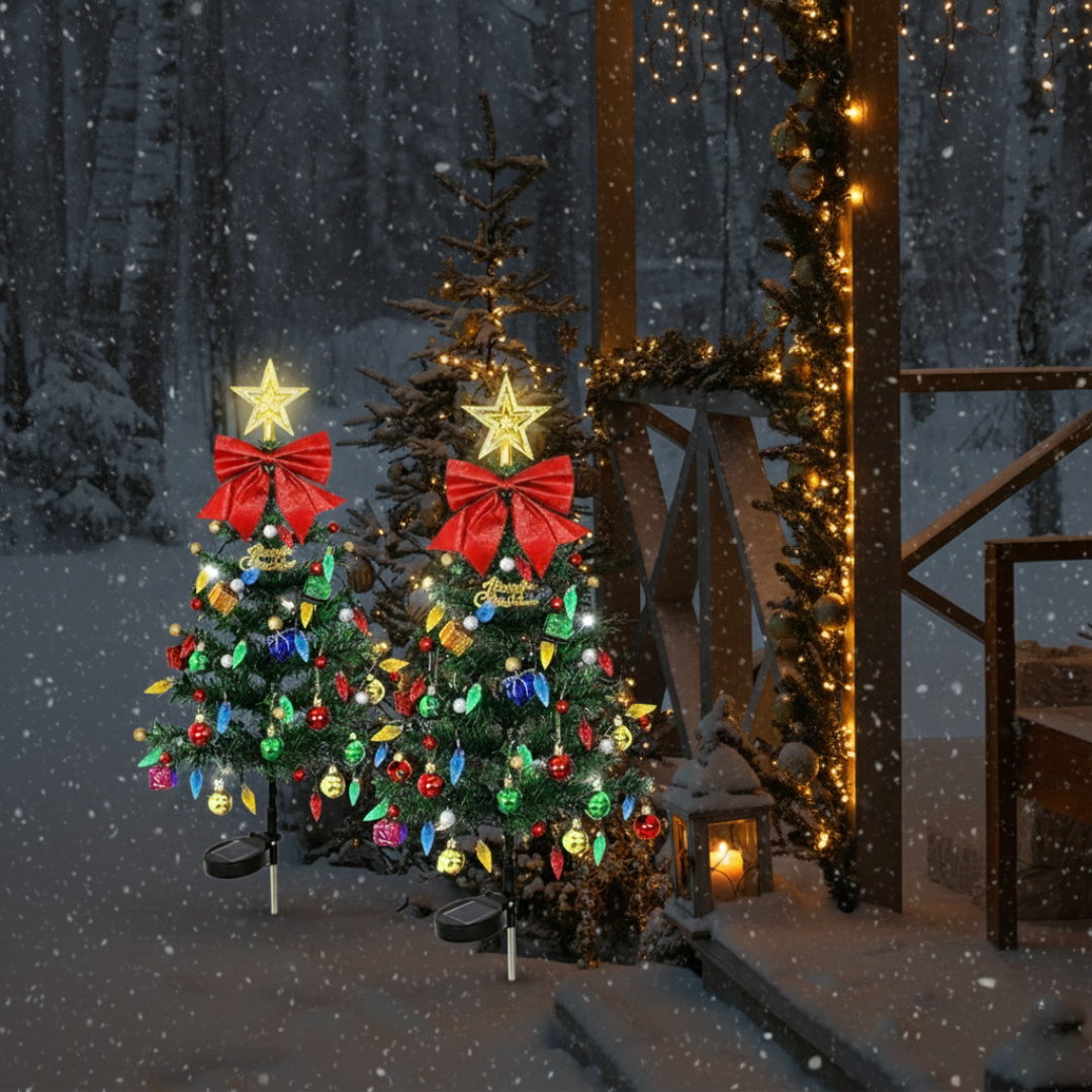 Two solar prelit outdoor Christmas trees with star toppers, red bows, and multicolor ornaments staked in snow beside a lit wooden structure at night
