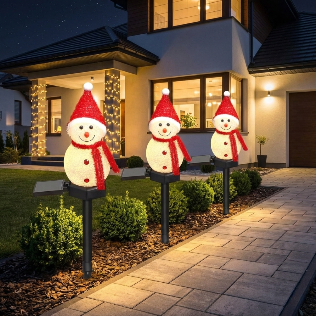 Three solar snowman pathway lights wearing red Santa hats and scarves staked along a garden border beside a stone walkway at night