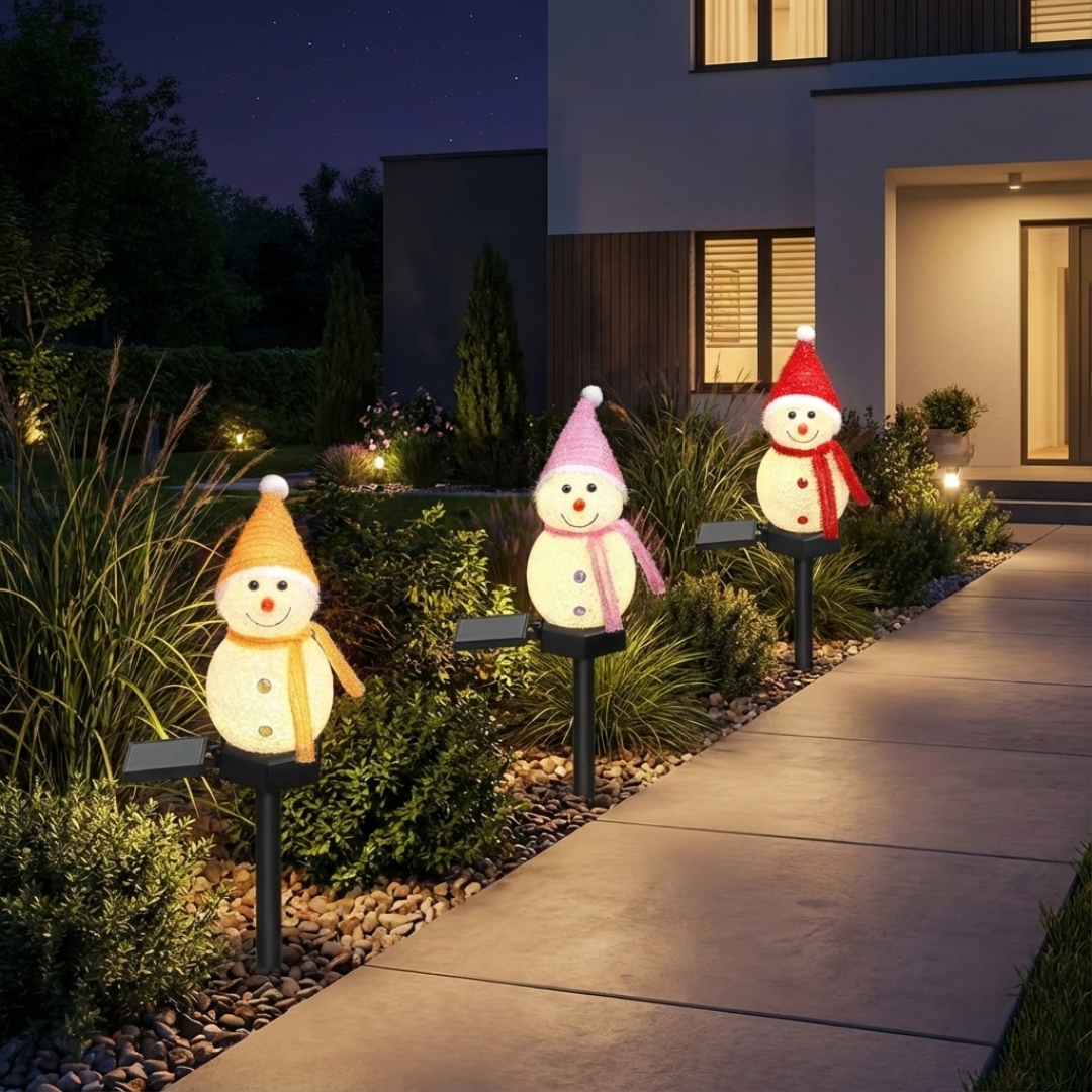 Three solar snowman pathway lights with yellow, pink, and red hats staked in a garden rock bed beside a paved walkway at night