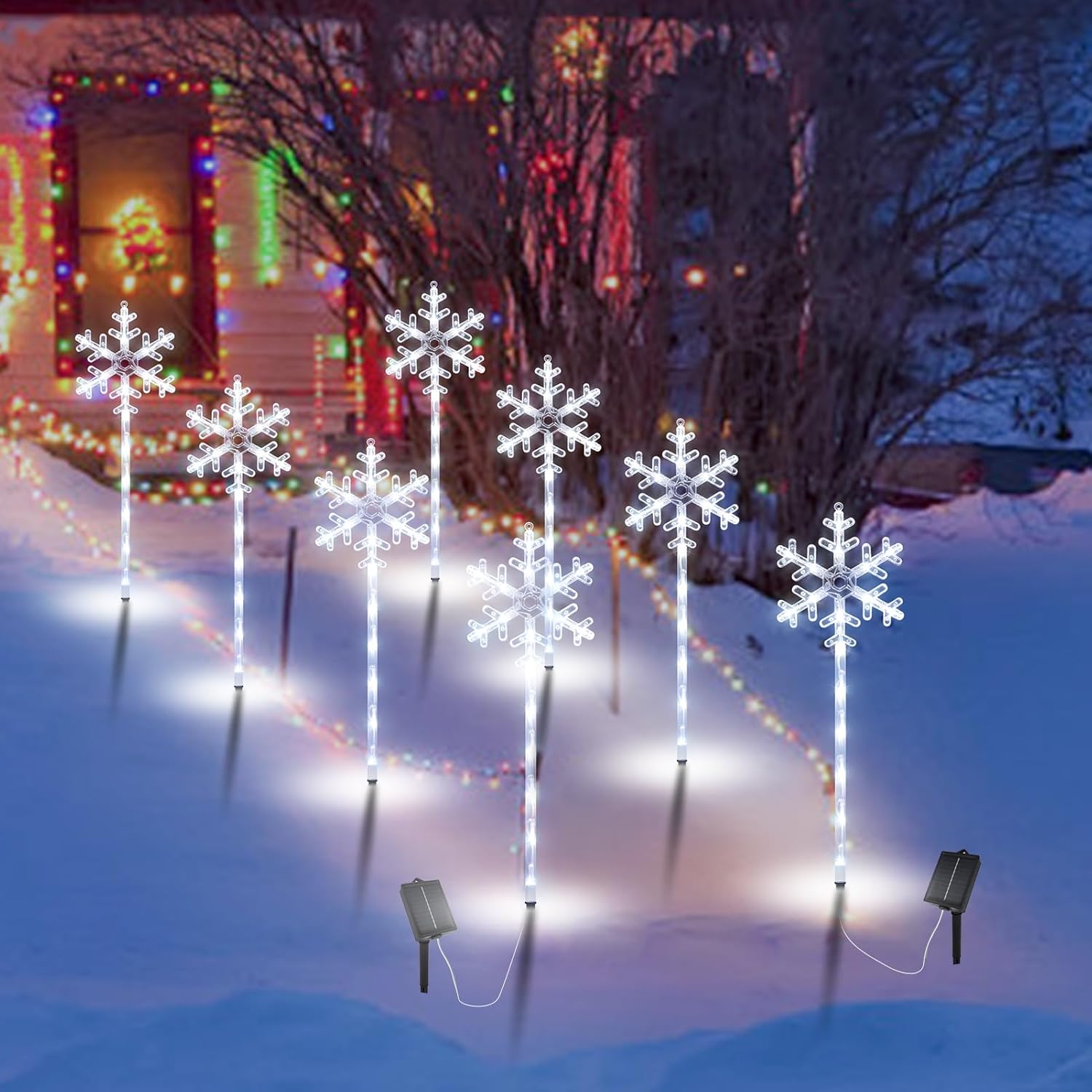 Eight solar snowflake pathway lights staked in deep snow at dusk, two solar panels visible, colorful holiday lights on house in background